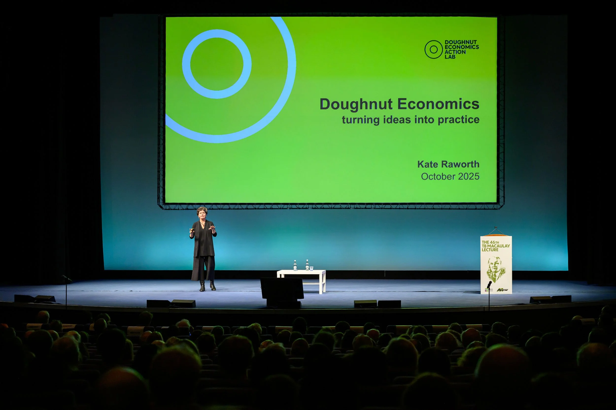 A woman gives a presentation on stage at a conference, with a large green slide displaying the title 'Doughnut Economics: turning ideas into practice' and the name 'Kate Raworth' and the date 'October 2025.' The audience is seated in front of the sta