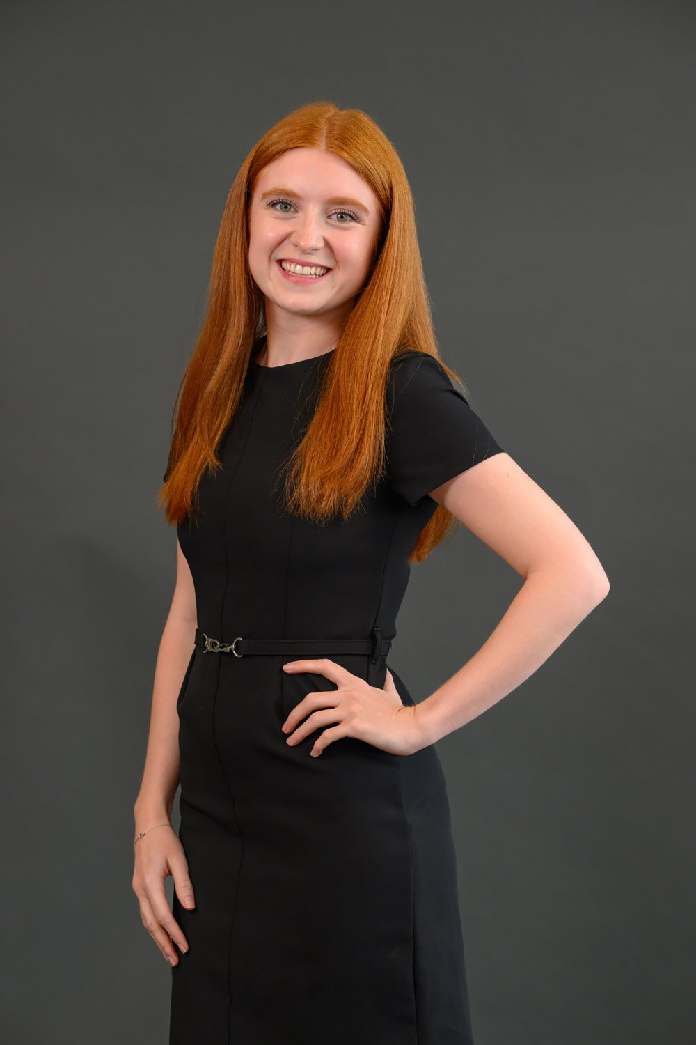 A woman with long red hair wearing a black dress and smiling at the camera against a plain gray background.
