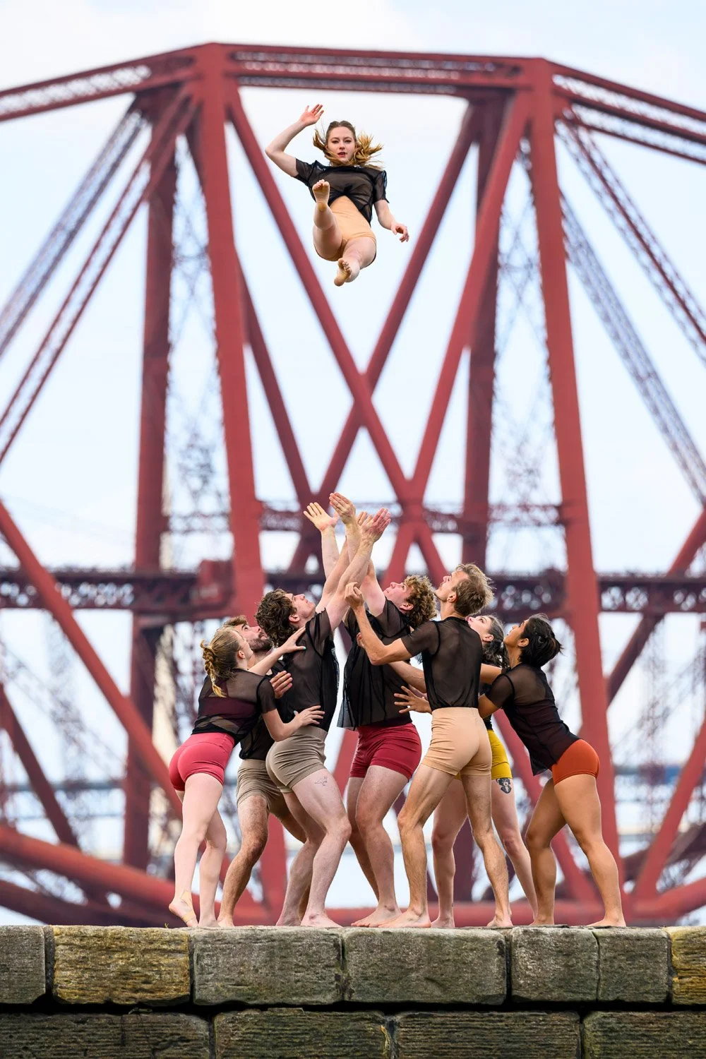 A group of dancers performs a coordinated lift and toss on a stone platform, with a red bridge structure in the background.