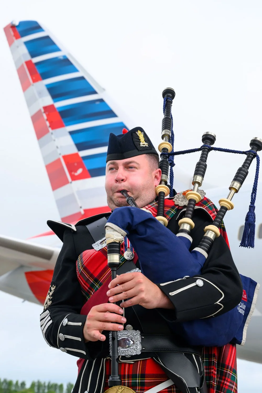 A man in Scottish Highland dress playing bagpipes at an airport with a large airplane tail in the background.