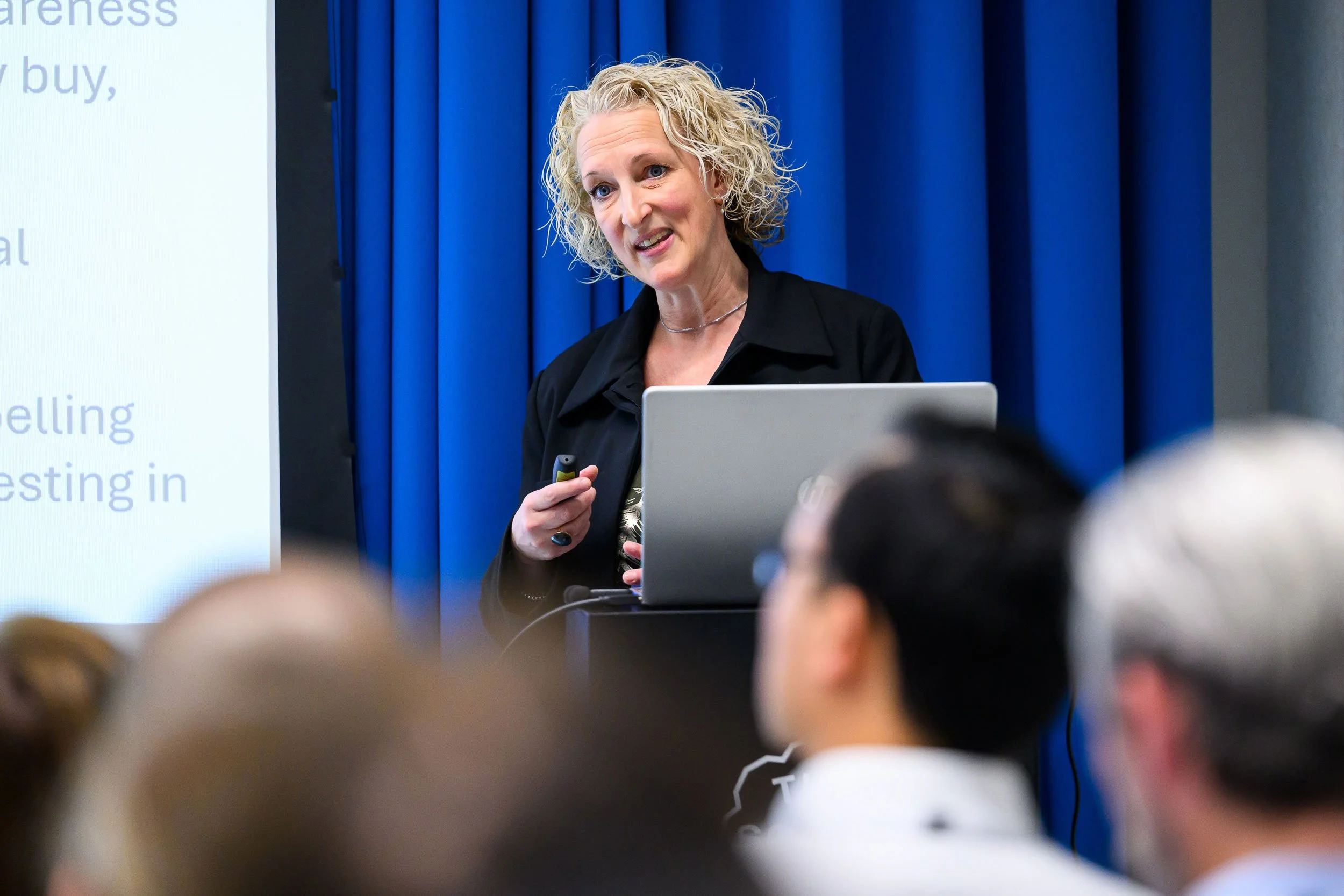 A woman with curly blonde hair giving a presentation in front of an audience, standing next to a large screen and a laptop, with blue curtains in the background.