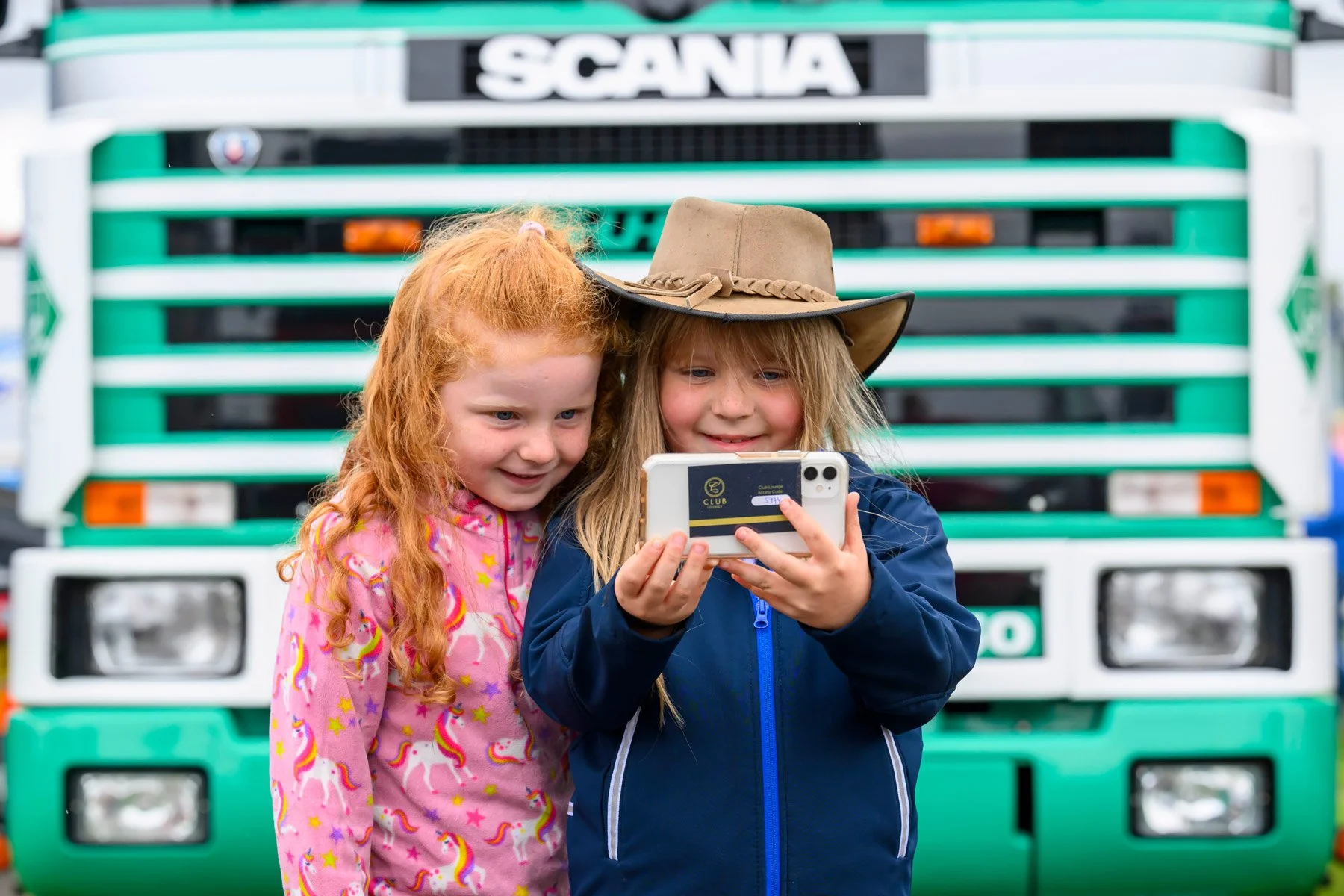 Two young girls with red and blonde hair smiling and looking at a smartphone in front of a large green and white truck.
