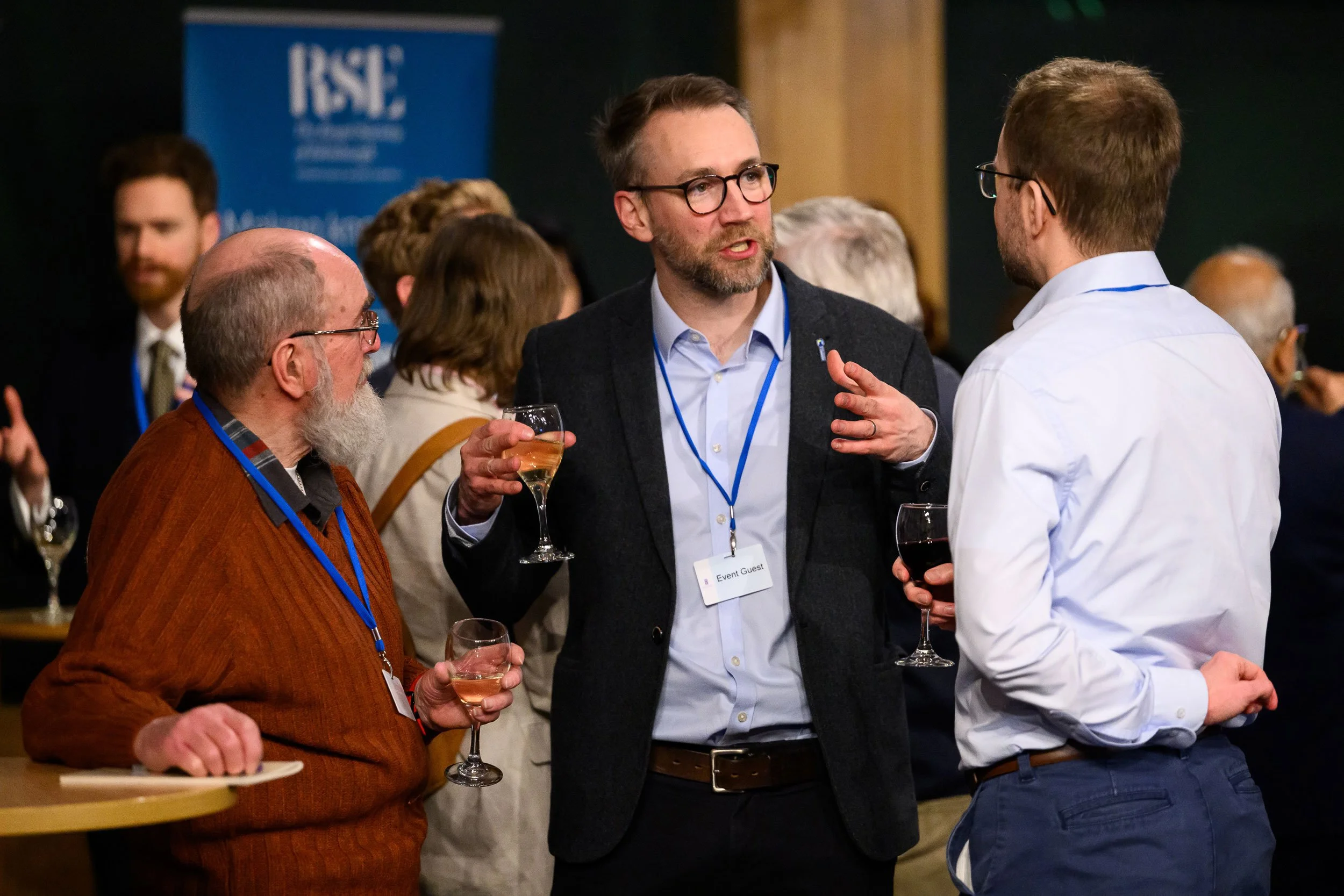 Group of people at a networking event, engaging in conversation, holding glasses of wine, with a blue conference banner in the background.