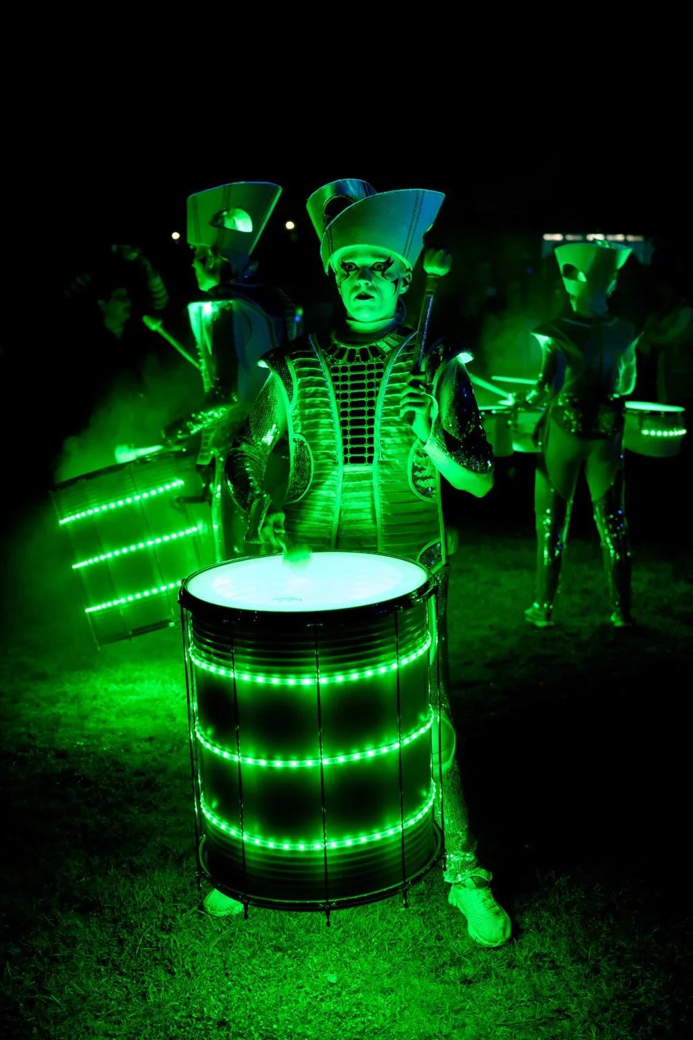 A performer dressed in a futuristic costume with glowing green lights, holding a large drum with illuminated bands, at a nighttime event with other performers in similar costumes in the background.