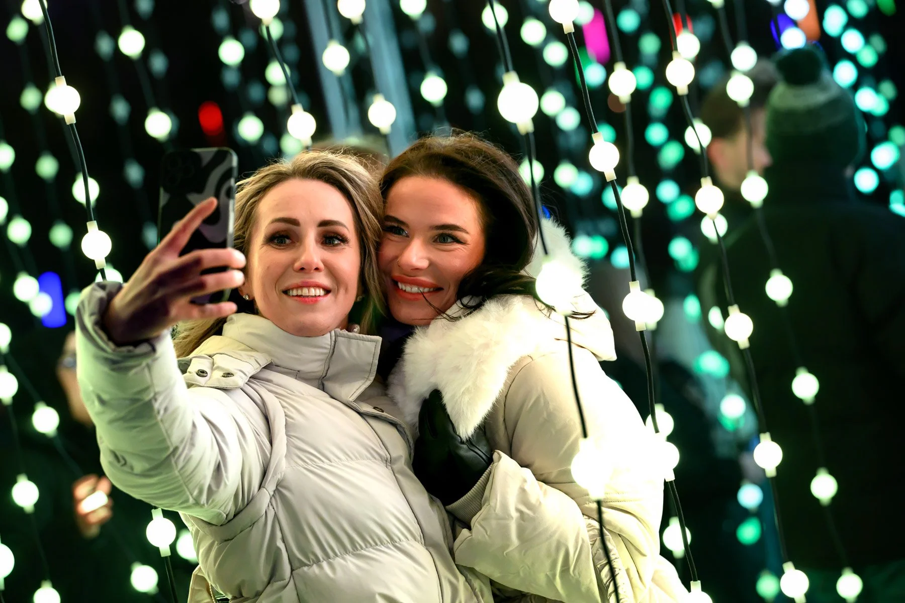 Two women taking a selfie in front of a vibrant, colorful light display, dressed warmly for winter.