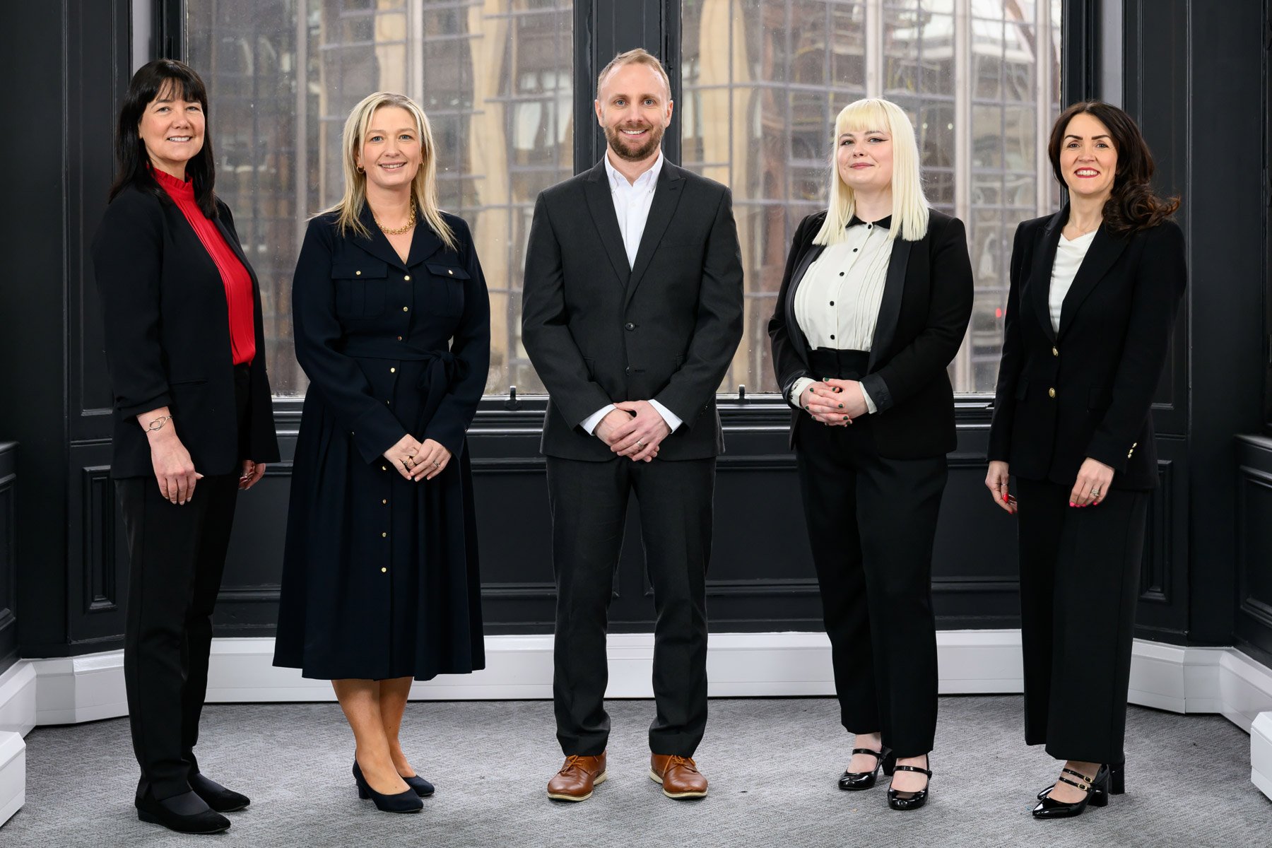 Group of five professionally dressed people standing in a room with large window behind them.
