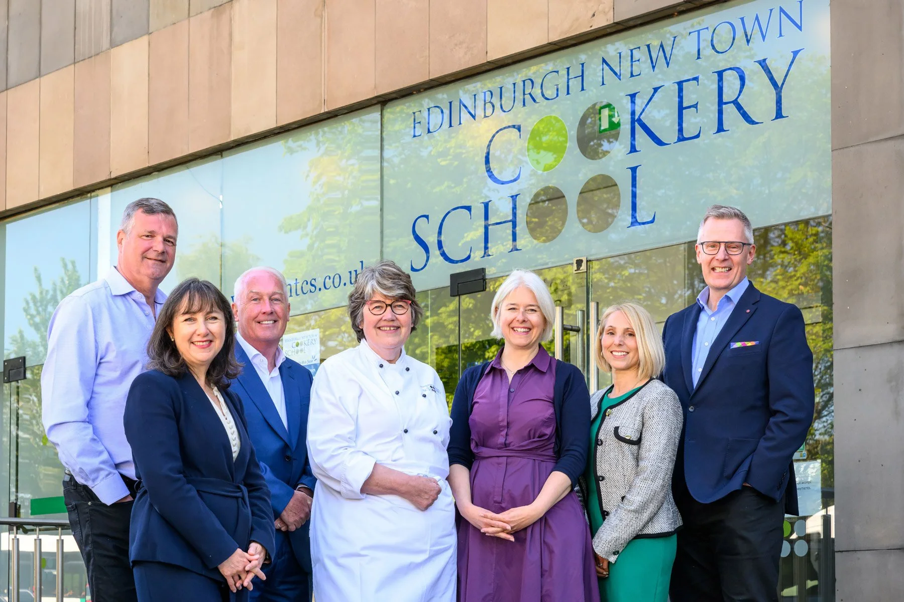Group of seven adults standing outside in front of the Edinburgh New Town Cookery School sign, smiling at the camera.