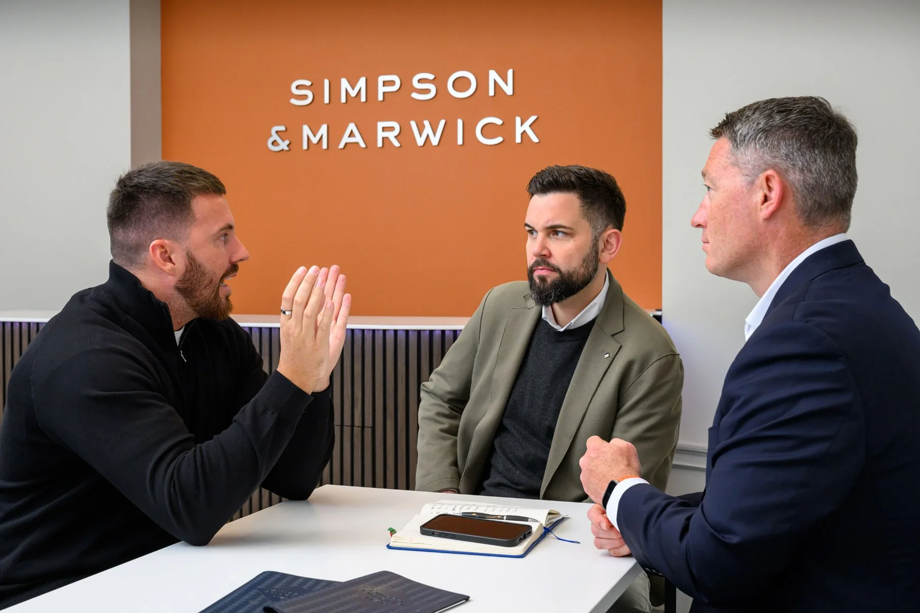 Three men having a serious discussion at a meeting table with a "SIMPSON & MARWICK" sign in the background.