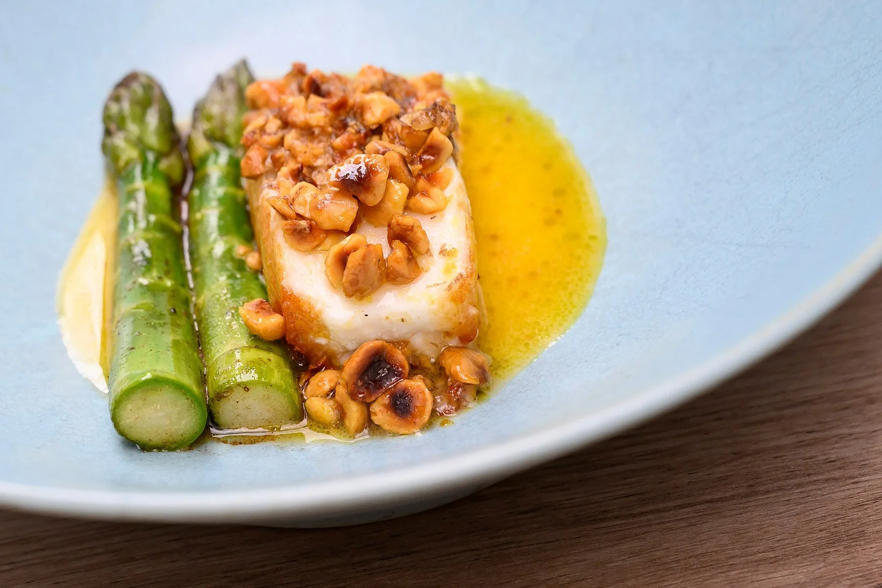 A white fish fillet topped with chopped nuts, served alongside grilled asparagus with a yellow sauce on a light blue plate.