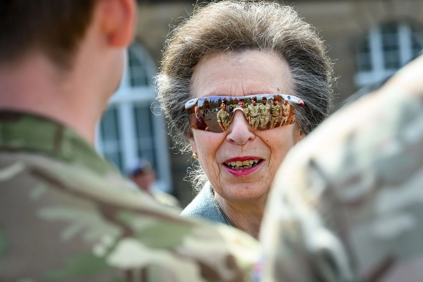 An elderly woman wearing reflective sunglasses with soldiers' reflection in the lenses, engaging in conversation with two soldiers in camouflage uniforms outdoors.