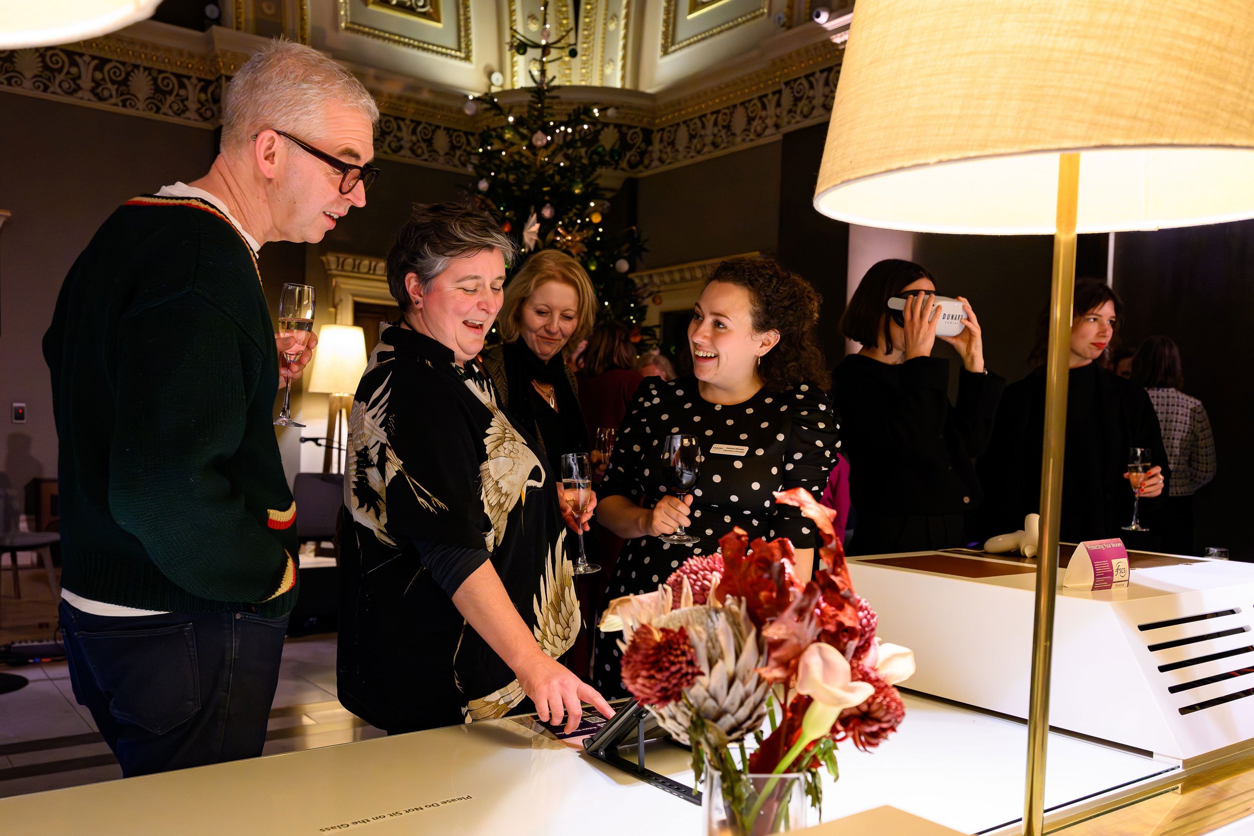 People gathered around a table at a holiday party, with a Christmas tree in the background, celebrating and enjoying drinks.