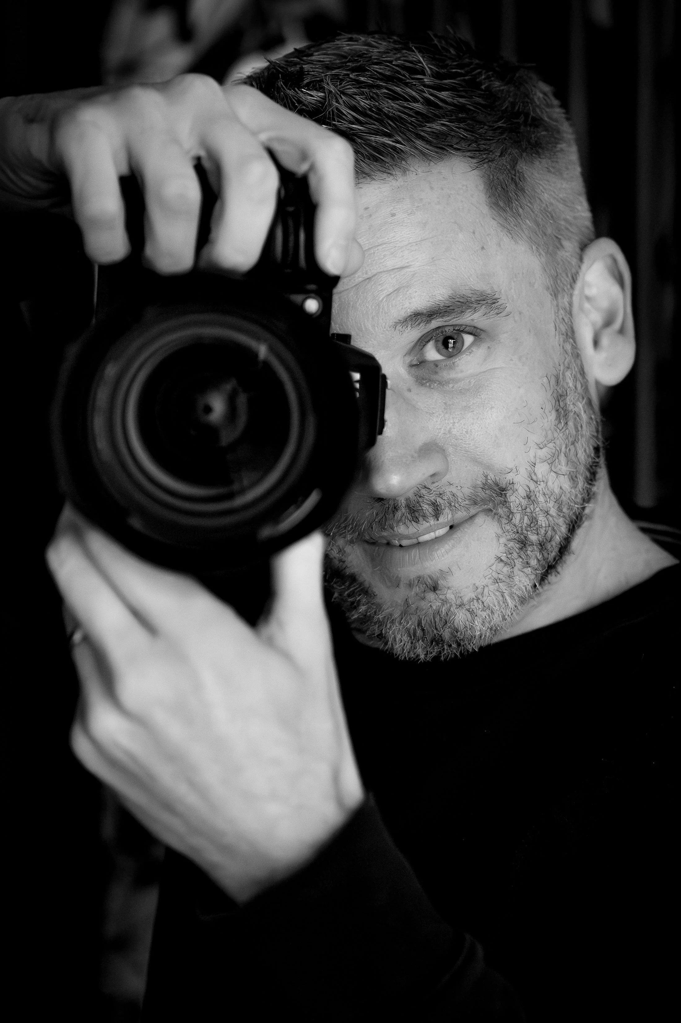 A man with a beard and short hair taking a selfie while holding a camera. The image is in black and white, and the man is smiling slightly.