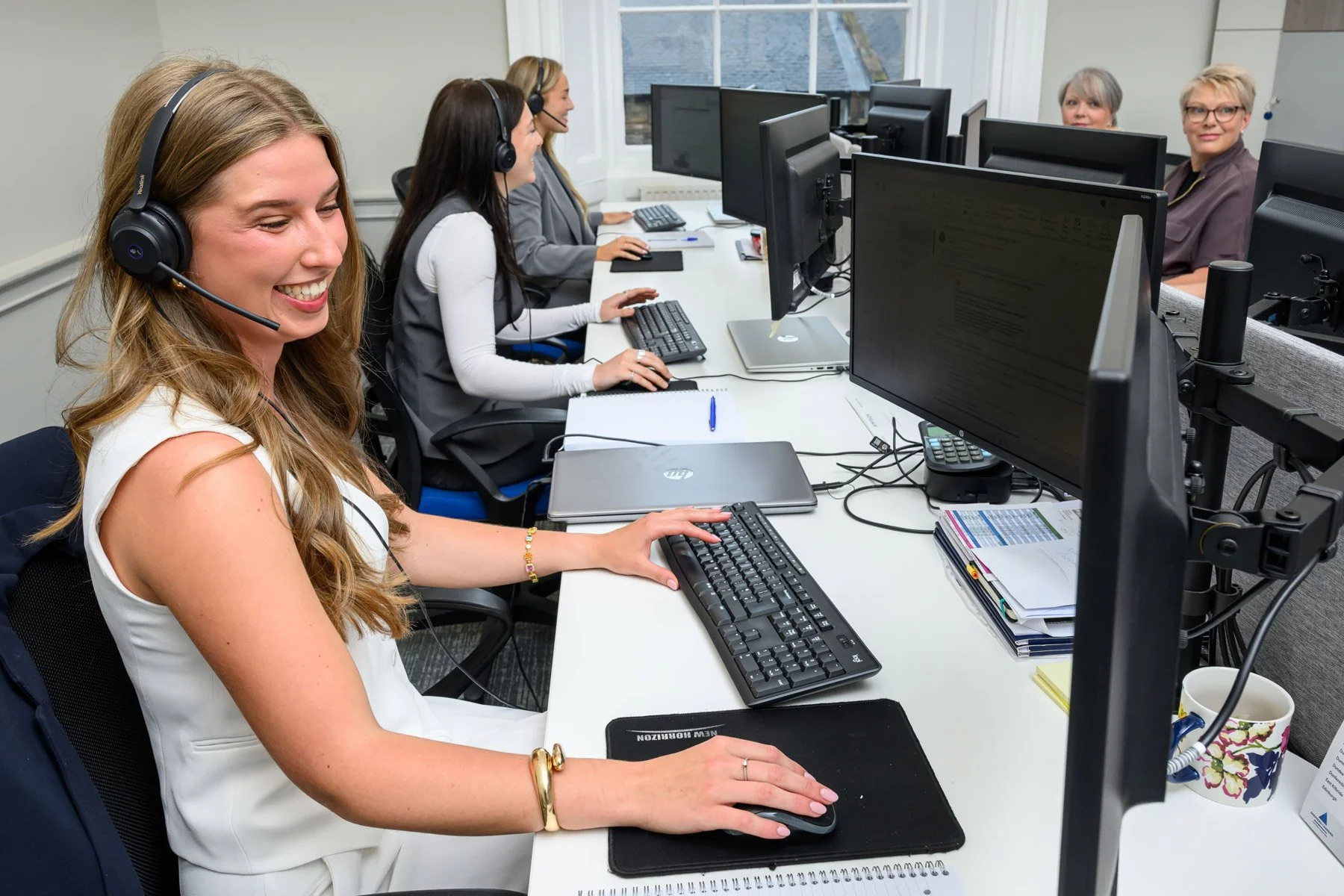 A group of women working at desks in an office, wearing headsets, using computers, with some smiling and others focused on their screens.