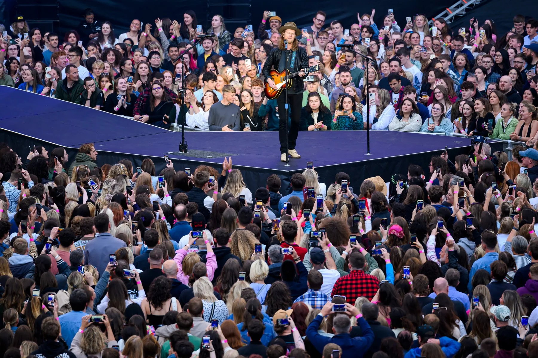 A musician with a cowboy hat and a guitar performs on stage in front of a large crowd holding up phones to record or take pictures, at an outdoor concert.