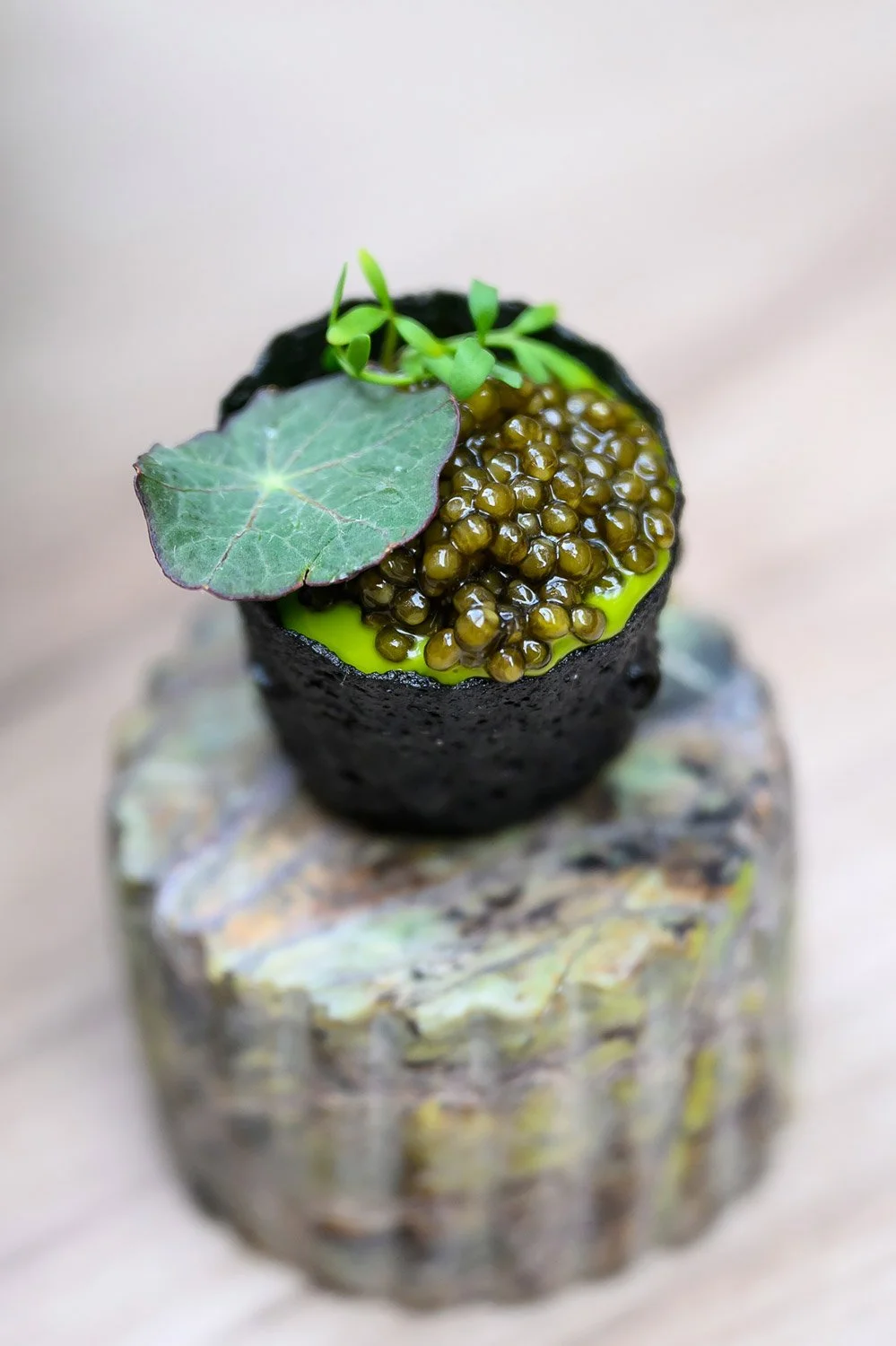 Close-up of a gourmet sushi roll topped with black caviar, a green leaf, and microgreens, on a textured wooden surface.