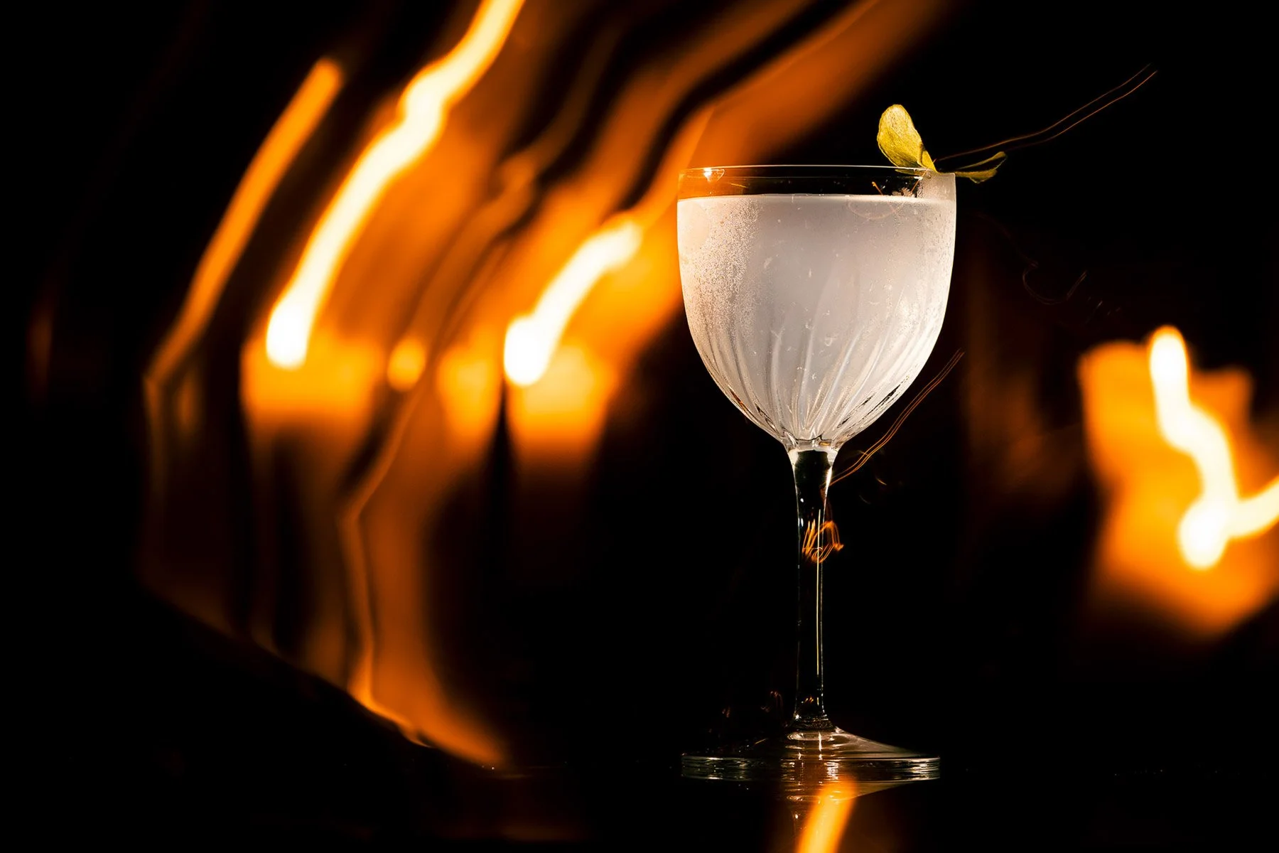 A close-up of a cocktail glass filled with a clear, frosted beverage, garnished with a small yellow flower, against a dark background with warm, blurred light streaks.