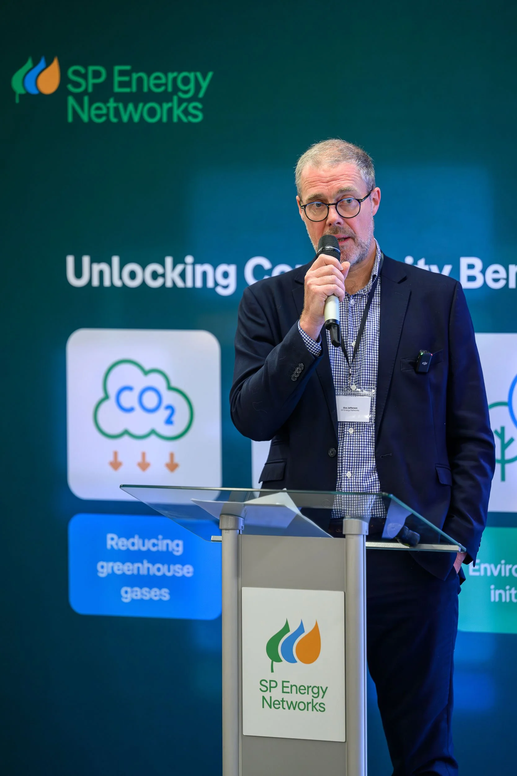 A man with glasses and a beard speaking into a microphone at a conference, standing behind a podium with SP Energy Networks logo, with a backdrop showing a presentation about reducing greenhouse gases and unlocking community benefits.