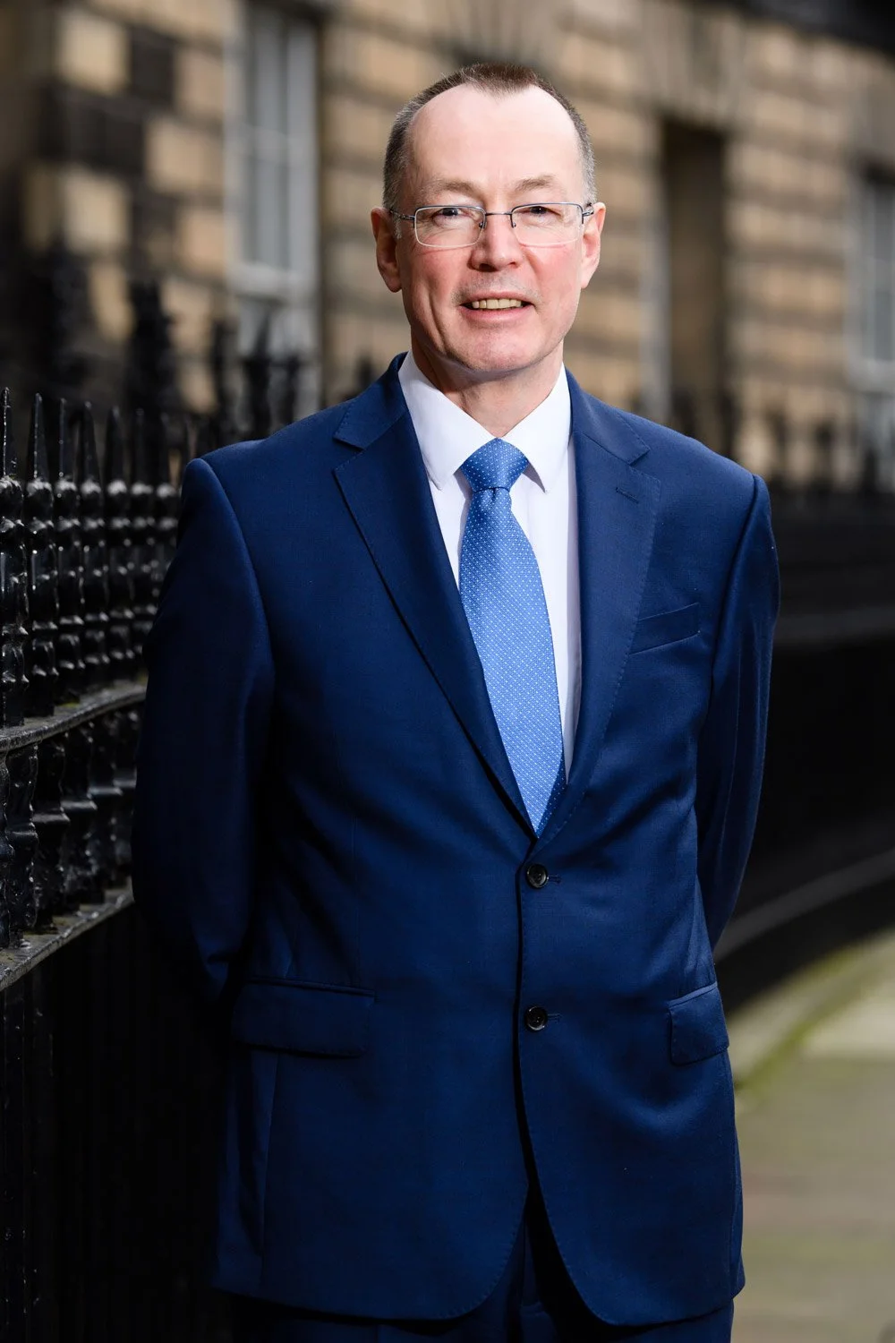 A man in a blue suit, white shirt, and blue tie standing outdoors near a black iron fence with a brick building in the background.