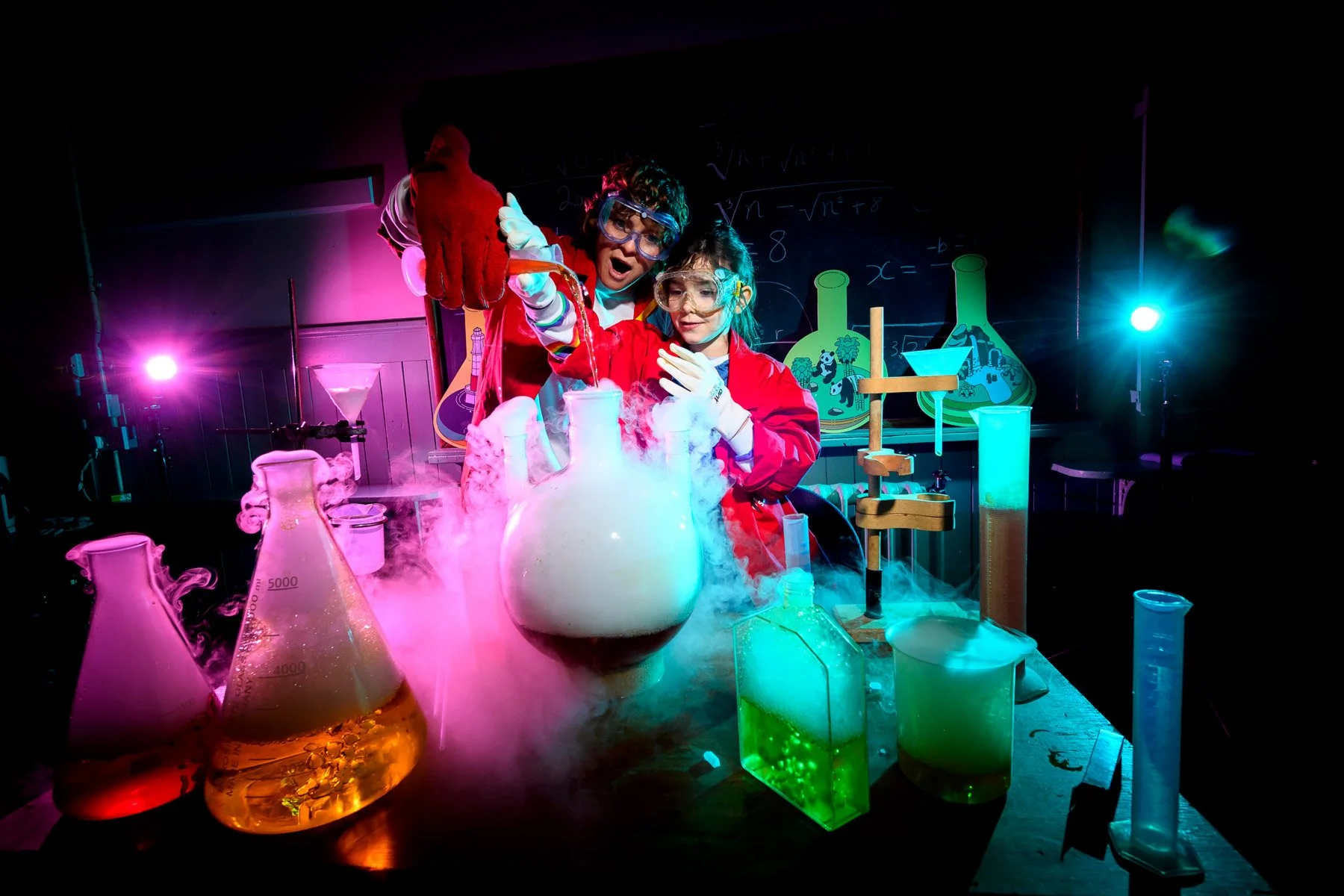 Two children in science lab coats and safety goggles conducting a chemistry experiment with colorful liquids in laboratory glassware surrounded by vapor and glowing lights.