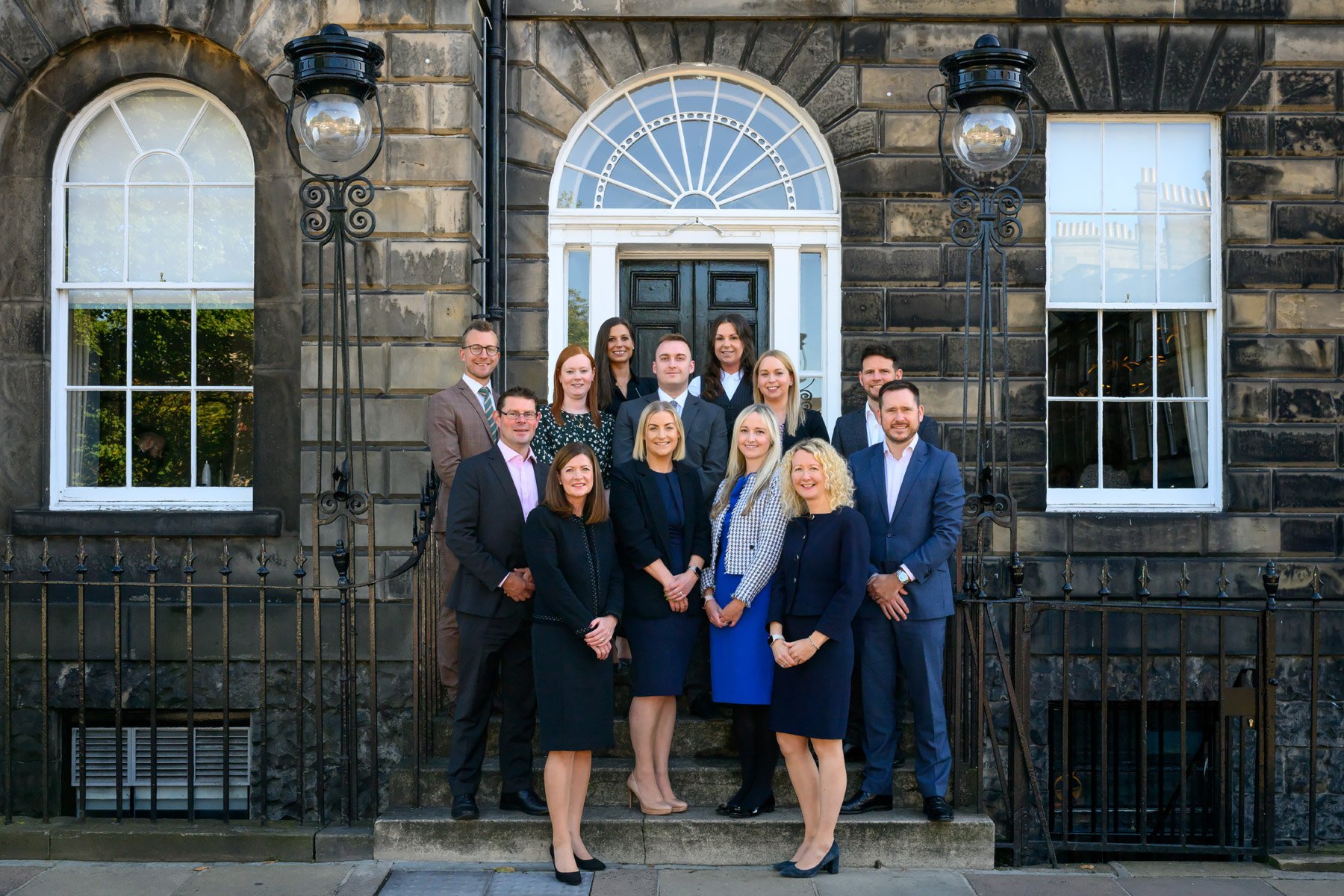 Group of professionally dressed men and women standing on steps in front of historic building with arched windows and black door.