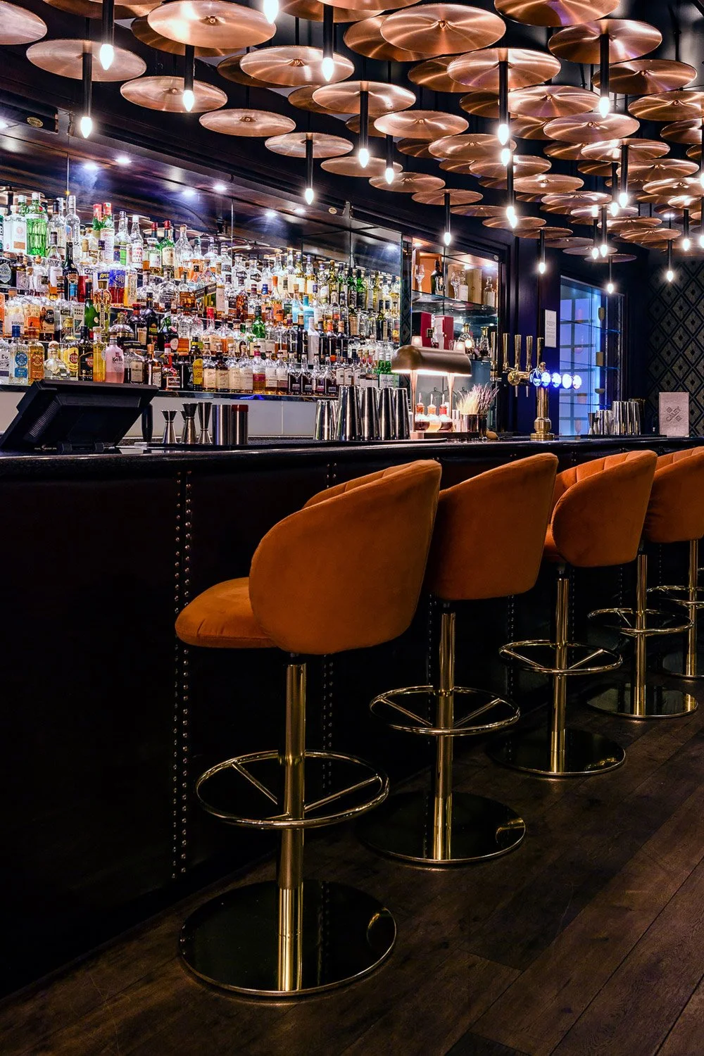 A stylish bar with a backlit liquor shelf filled with various bottles, copper pendant lights hanging from the ceiling, a dark bar counter with orange velvet bar stools, and beer taps.