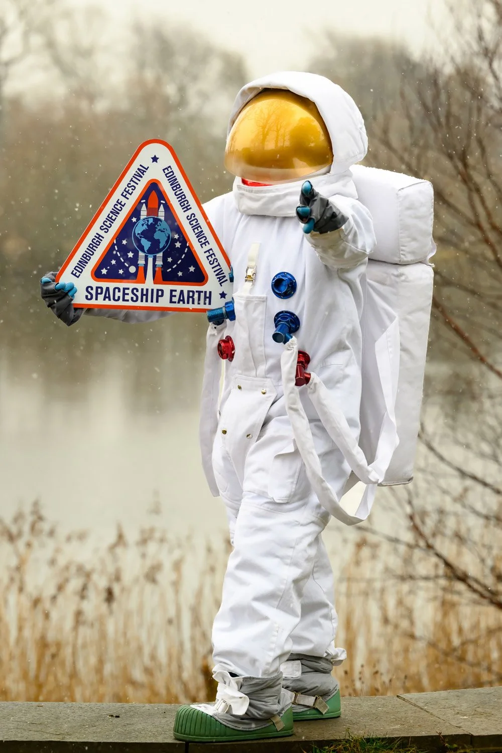 Person in a white astronaut suit holding a colorful badge that reads 'Spaceship Earth' and 'Edinburgh Science Festival'. The background features a natural outdoor scene with trees and a body of water.