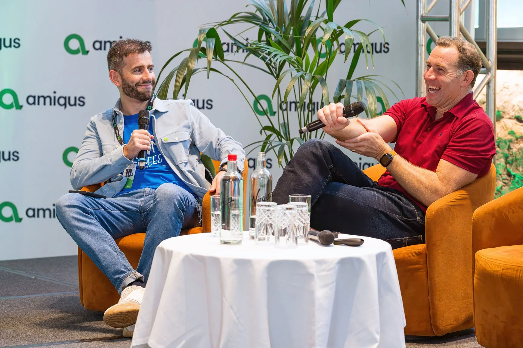 Two men sitting on orange chairs having a conversation on stage with microphones, surrounded by glasses and bottles of water, a small round table, and a backdrop with the logo 'amiqus', with a green plant nearby.