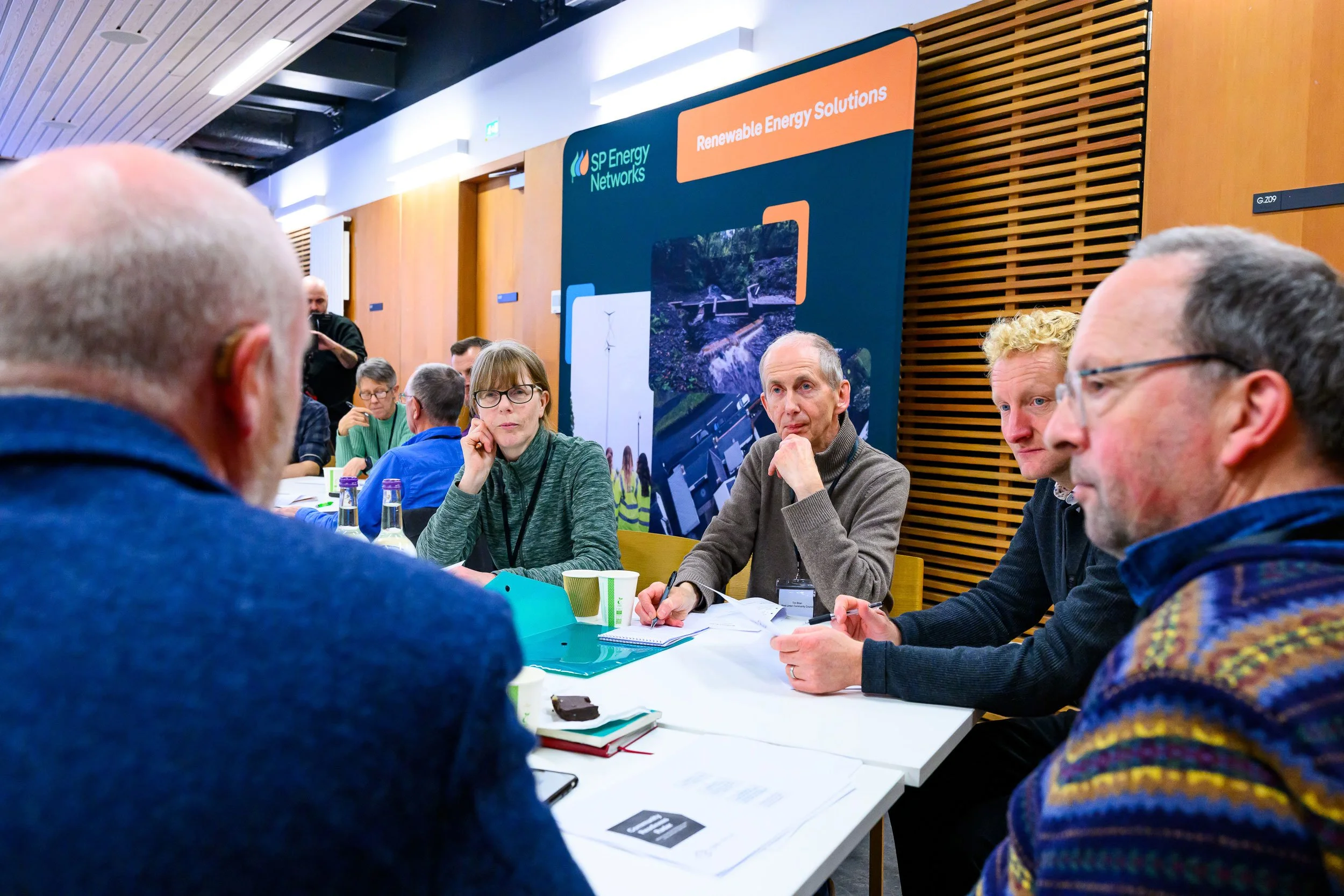 A group of people gathered around a table at a conference, engaging in discussion. In the background, a banner displays the logo of SP Energy Networks and the words "Renewable Energy Solutions."