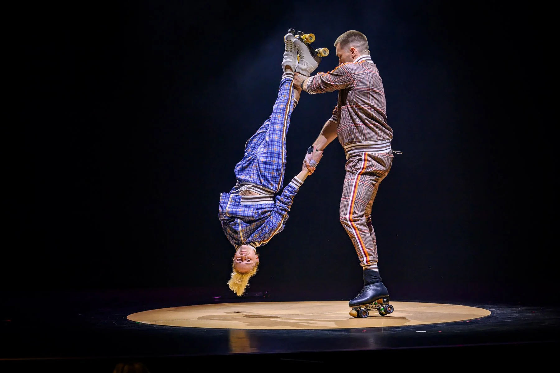 Two skaters on a stage, one is hanging upside down and holding the other's hand, while standing on roller skates; both are dressed in colorful outfits with plaid patterns.