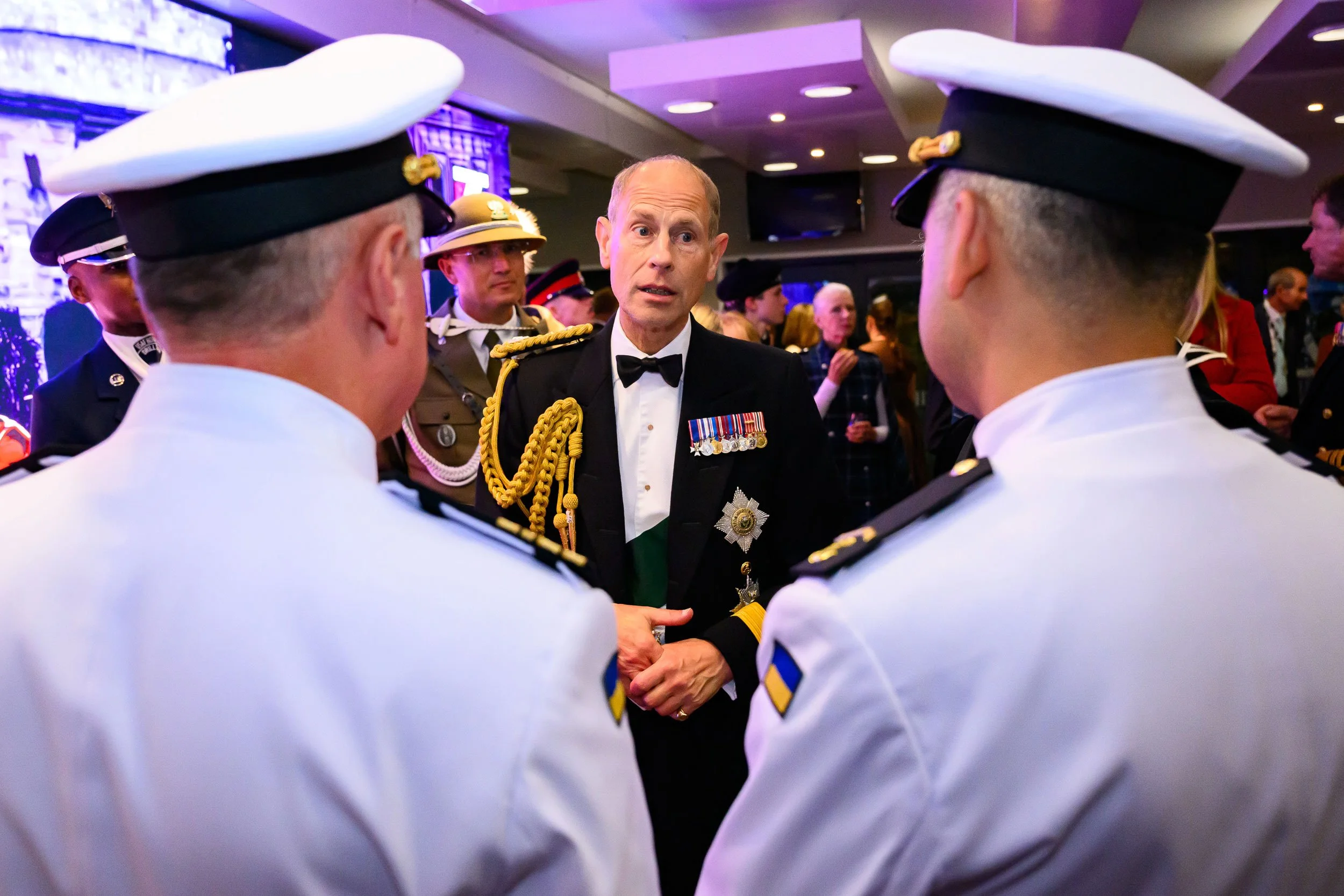 A group of military officers in uniform having a conversation at a formal event, with a man in a tuxedo in the center.