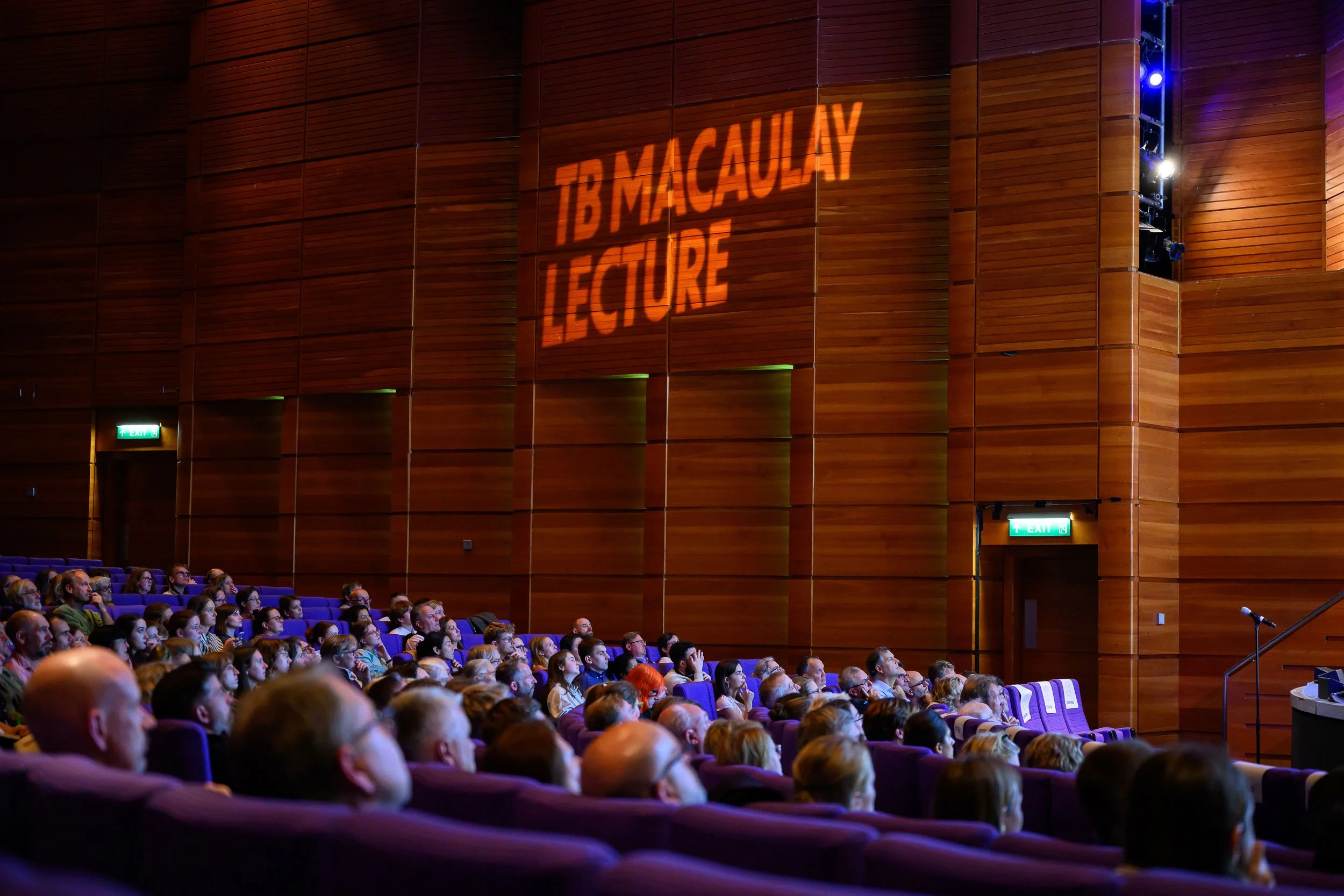 Audience listening at TB Macaulay Lecture in a large auditorium with wooden walls and purple seats