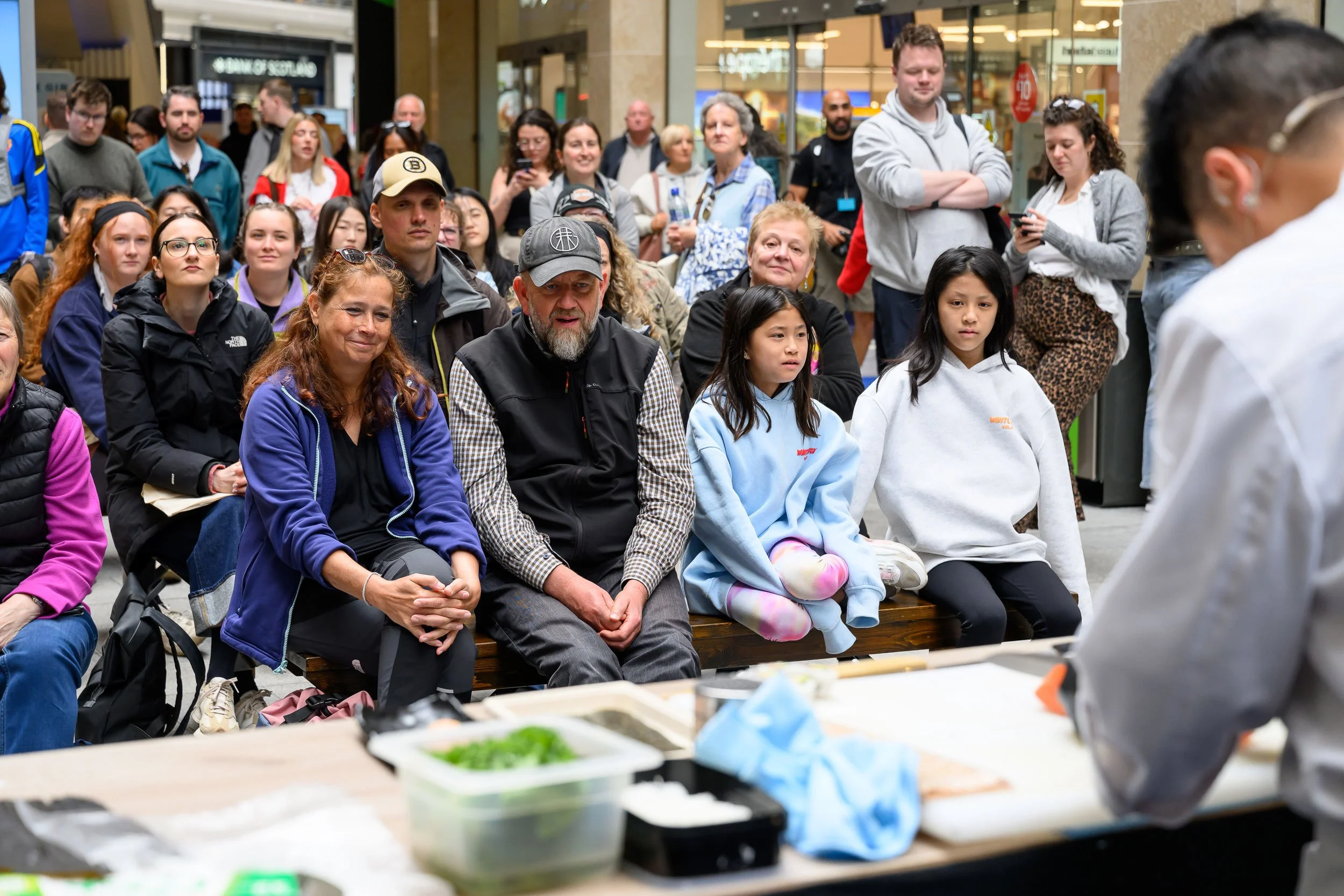 People sitting on benches in a shopping mall watching a demonstration or cooking class being conducted by a person in a chef's coat. There are various food ingredients and utensils on the table in front of the chef.