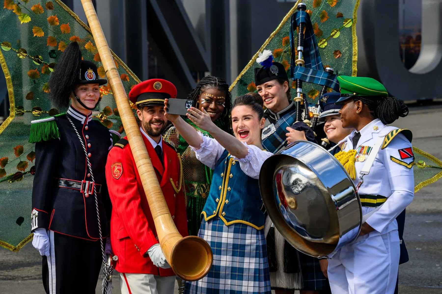 A group of six diverse people in colorful costumes and medals, posing for a selfie in front of a decorated parade float with a green canopy and gold accents.