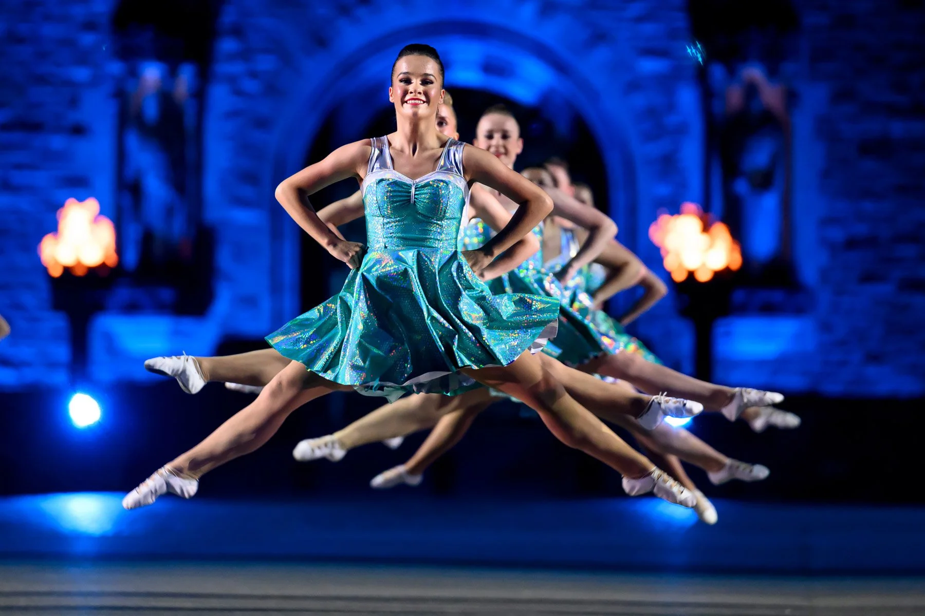 Group of ballerinas in iridescent blue costumes performing a synchronized jump on stage with a blue-lit stone castle backdrop.