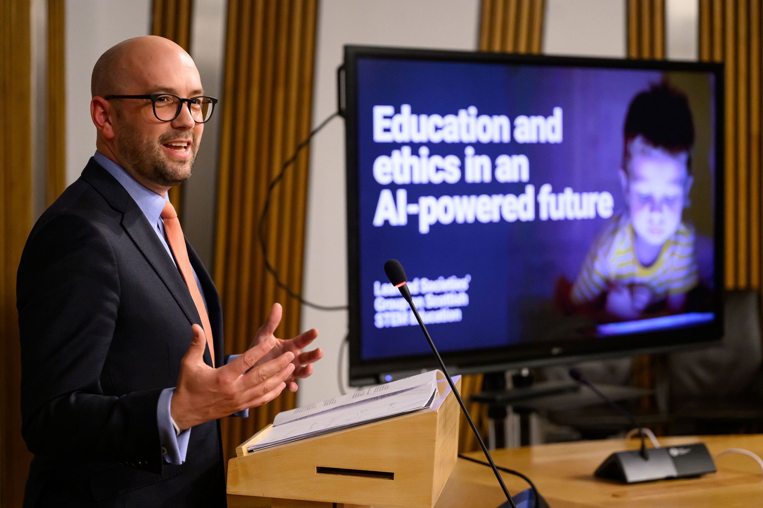 A man in a suit and glasses giving a presentation at a podium, with a large screen behind him displaying the title 'Education and ethics in an AI-powered future' and an image of a young child.