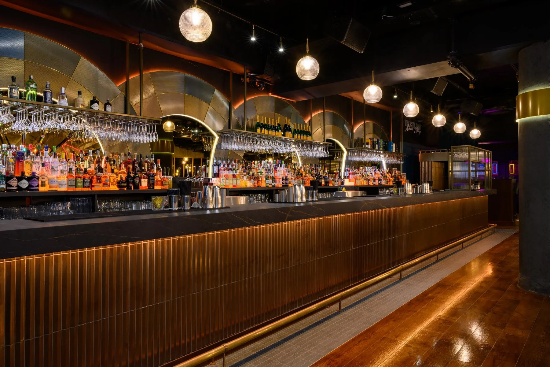 A modern bar with a dark color scheme, illuminated by spherical pendant lights. It features a long counter with a variety of liquor bottles, glasses, and bar tools, and a reflective mirror with an artistic design in the background.