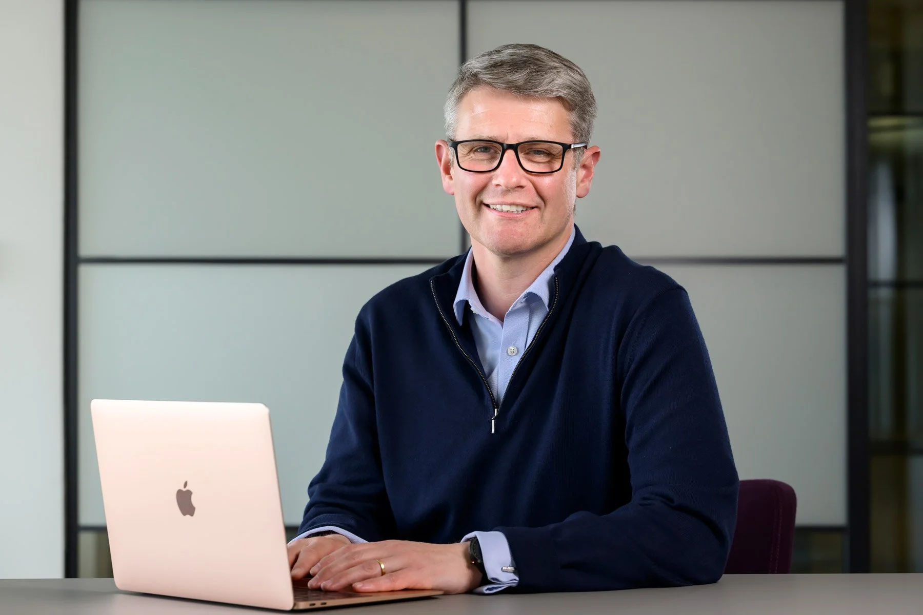 A man with glasses sitting at a desk using a silver MacBook laptop in an office environment.