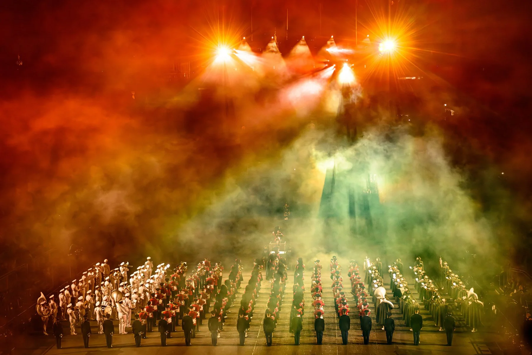 A large group of marching band members in uniform, standing in formation on a field, with colorful stage lights and smoke effects overhead.