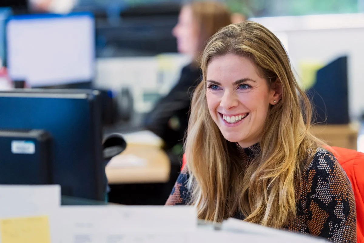 A smiling woman with long blonde hair working at a computer in an office environment.