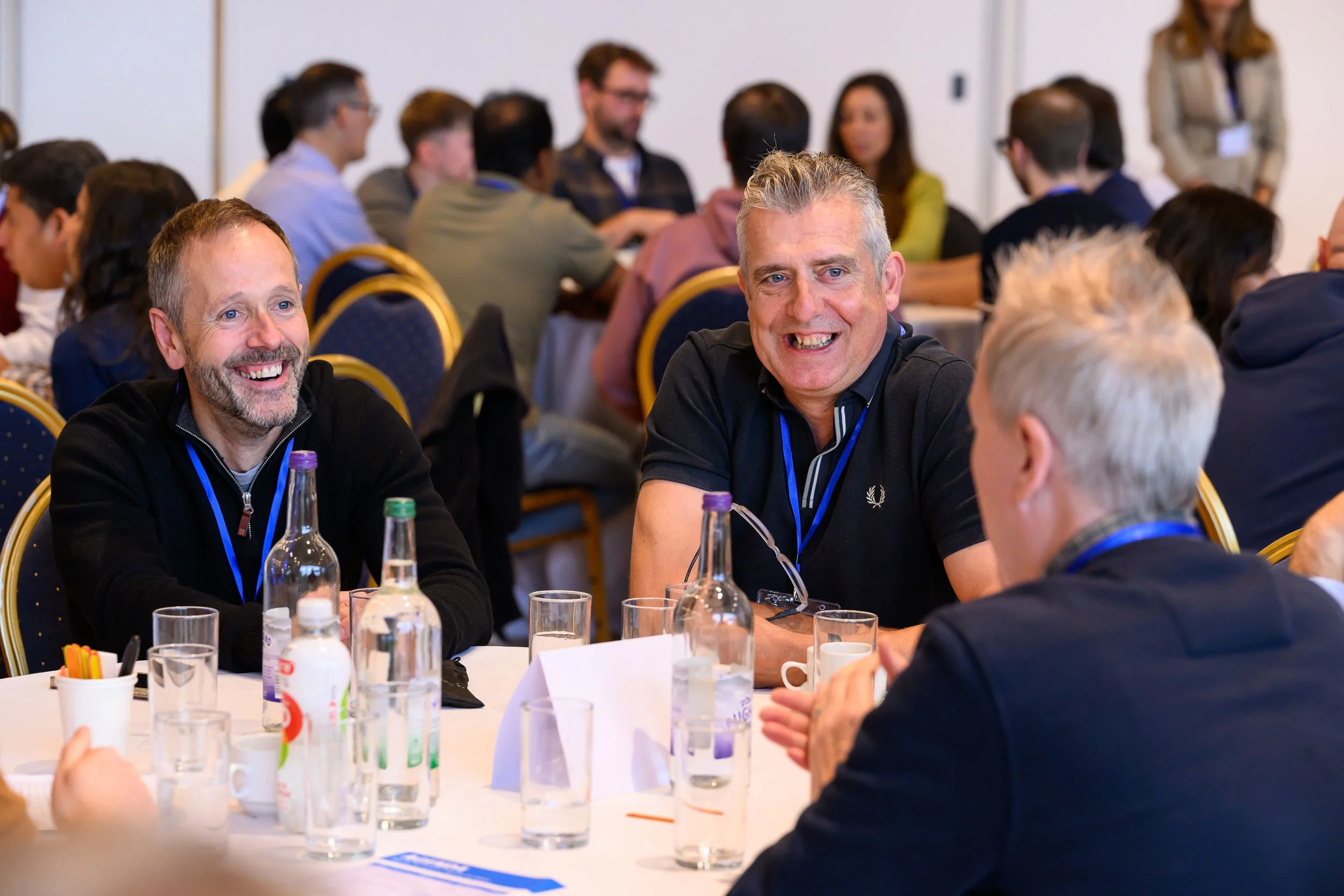 Three men are sitting at a round table, engaged in conversation and smiling, in a crowded conference room.