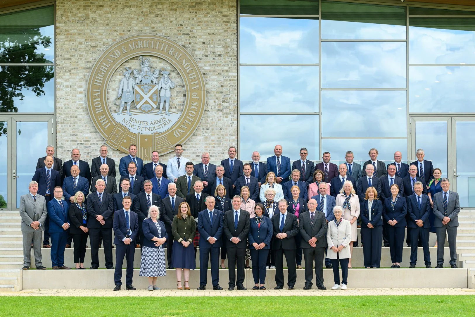 Group of people posed for a photo outside a building with a large emblem on the wall, representing the Royal Highland & Agricultural Society of Scotland.