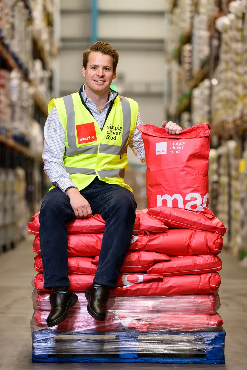 A man sitting on wrapped pallets of red bags with the Macphie and Simply Clever Food logos in a warehouse.