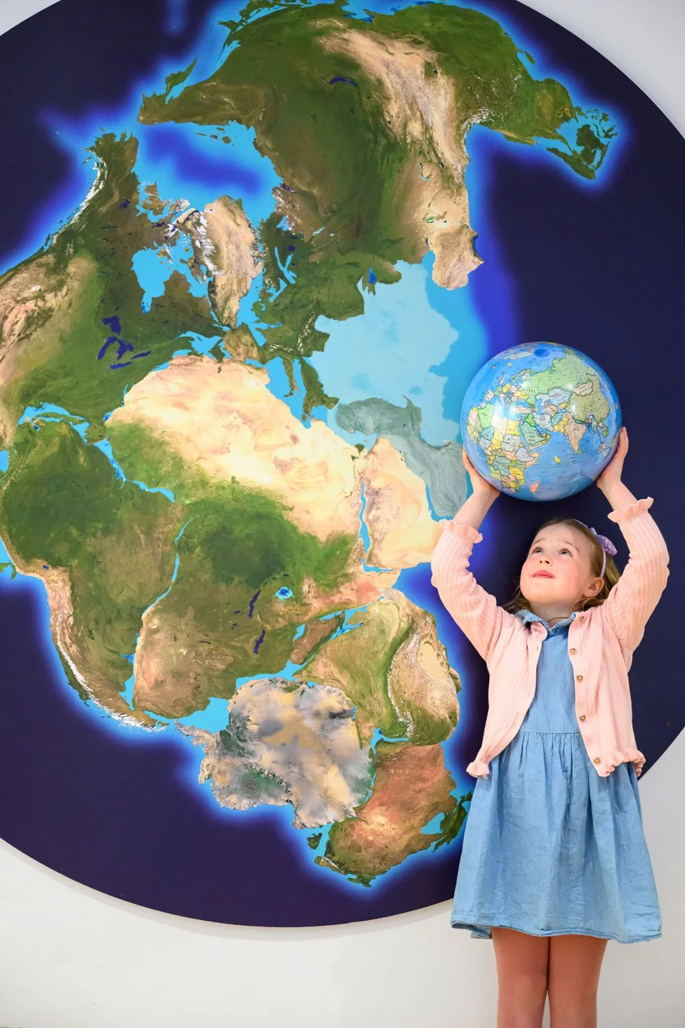 A young girl holding a globe above her head, standing in front of a large map of the world on the wall.