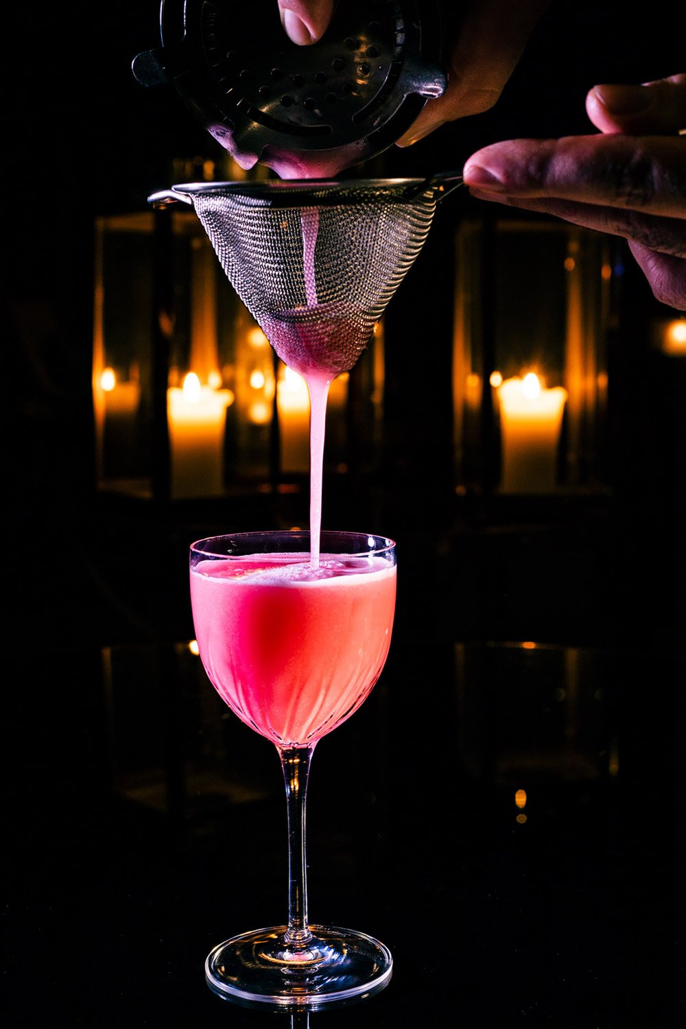 Pink cocktail being strained into a glass through a mesh strainer, with lit candles in the background.