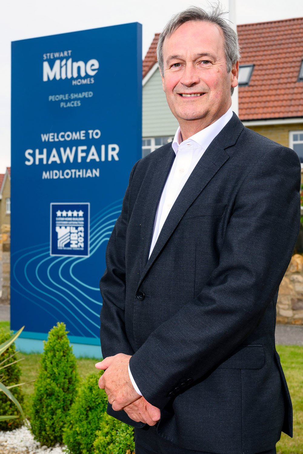 A man in a suit standing outdoors in front of a blue sign that reads "Welcome to Shawfair Midlothian," with a logo and some decorative lines, with houses in the background.
