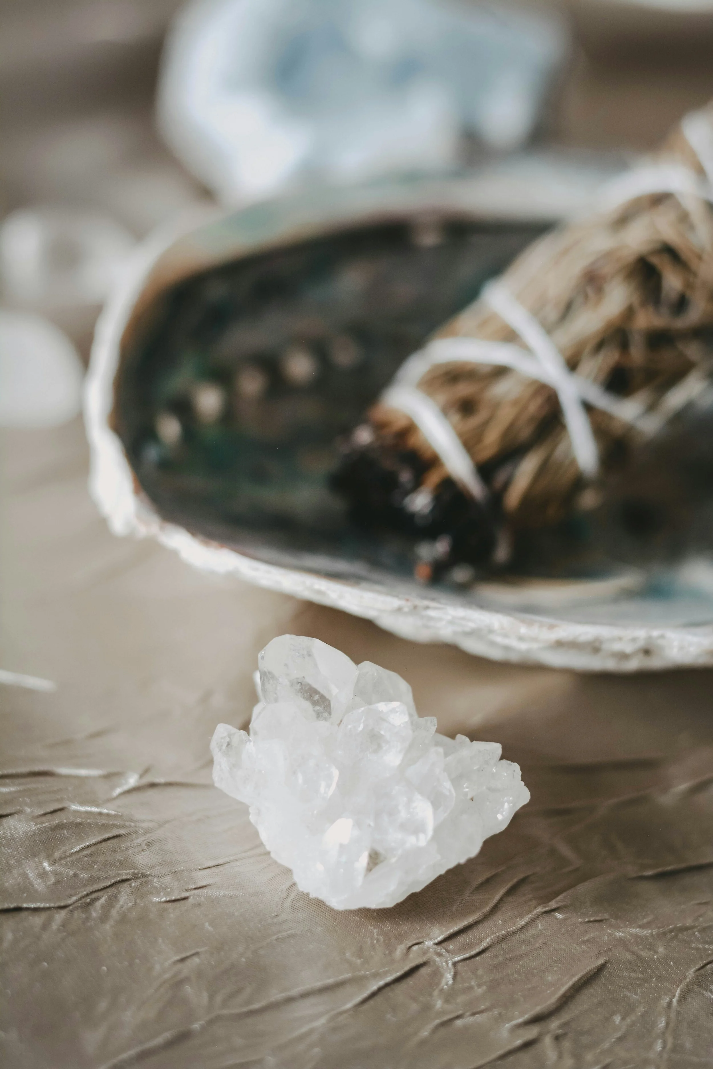 Close-up of a piece of white crystalline salt crystal in the foreground and a conch shell with a bundle of dried herbs secured with white string resting inside on a textured beige surface.