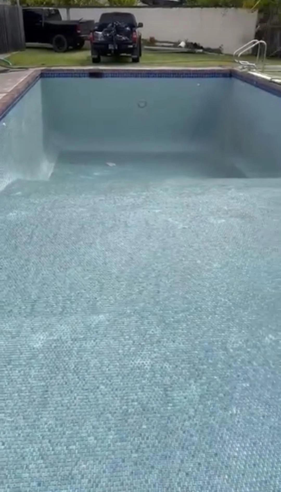 Empty swimming pool with a textured surface, with a ladder on the right side, a boat and vehicles in the background.