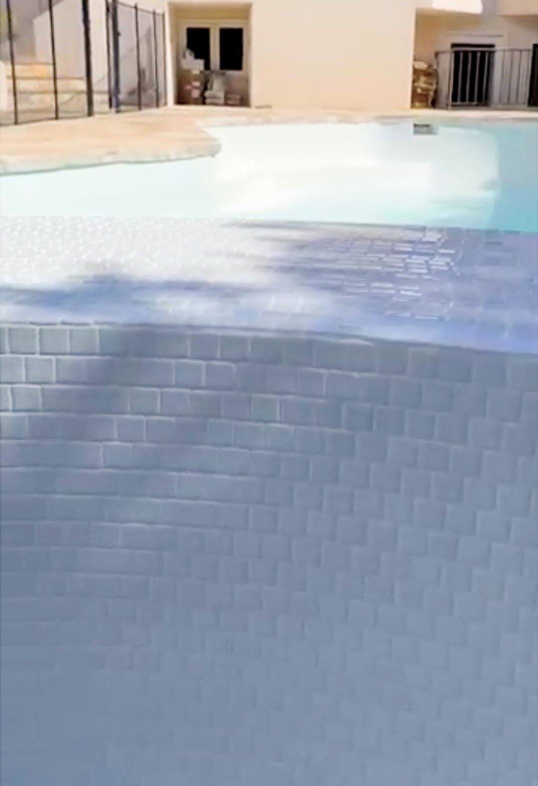 Close-up of a swimming pool with gray brick tile border and light blue water, with a beige patio around it.