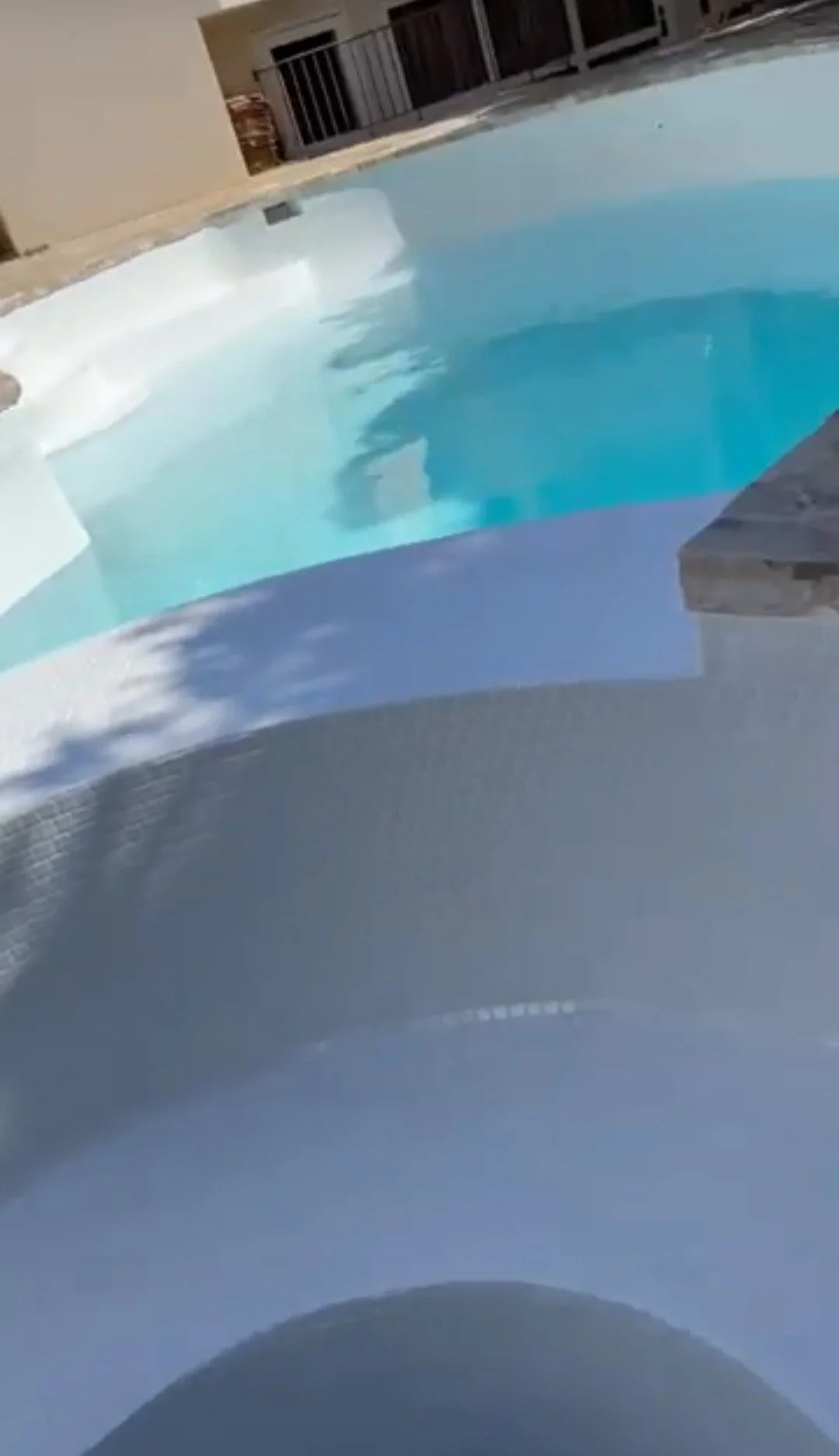 Swimming pool with blue water and a white edge, surrounded by a beige deck and a building with a balcony.