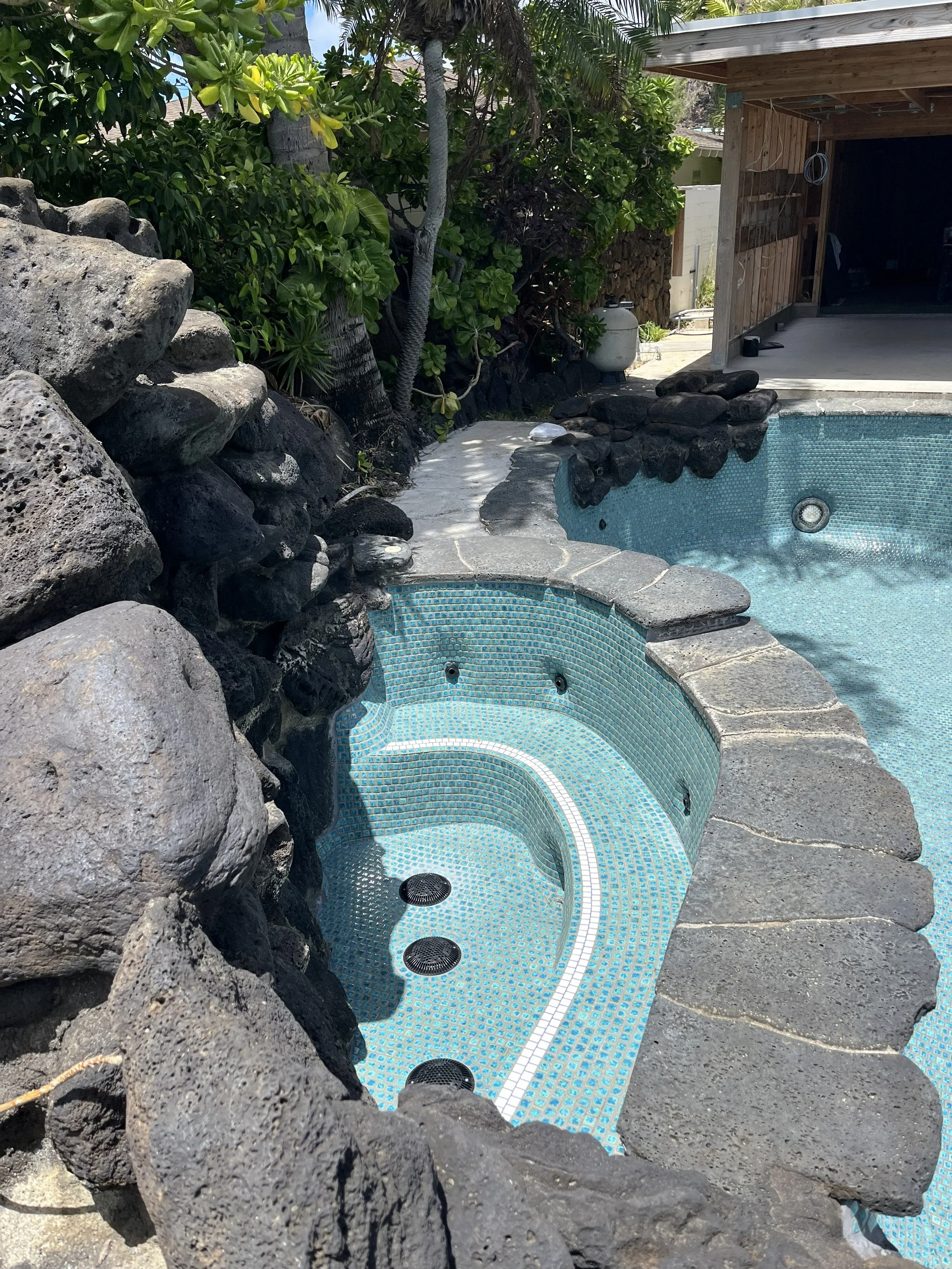 Empty hot tub with blue mosaic tiles surrounded by rocks, plants, and trees in an outdoor backyard setting.